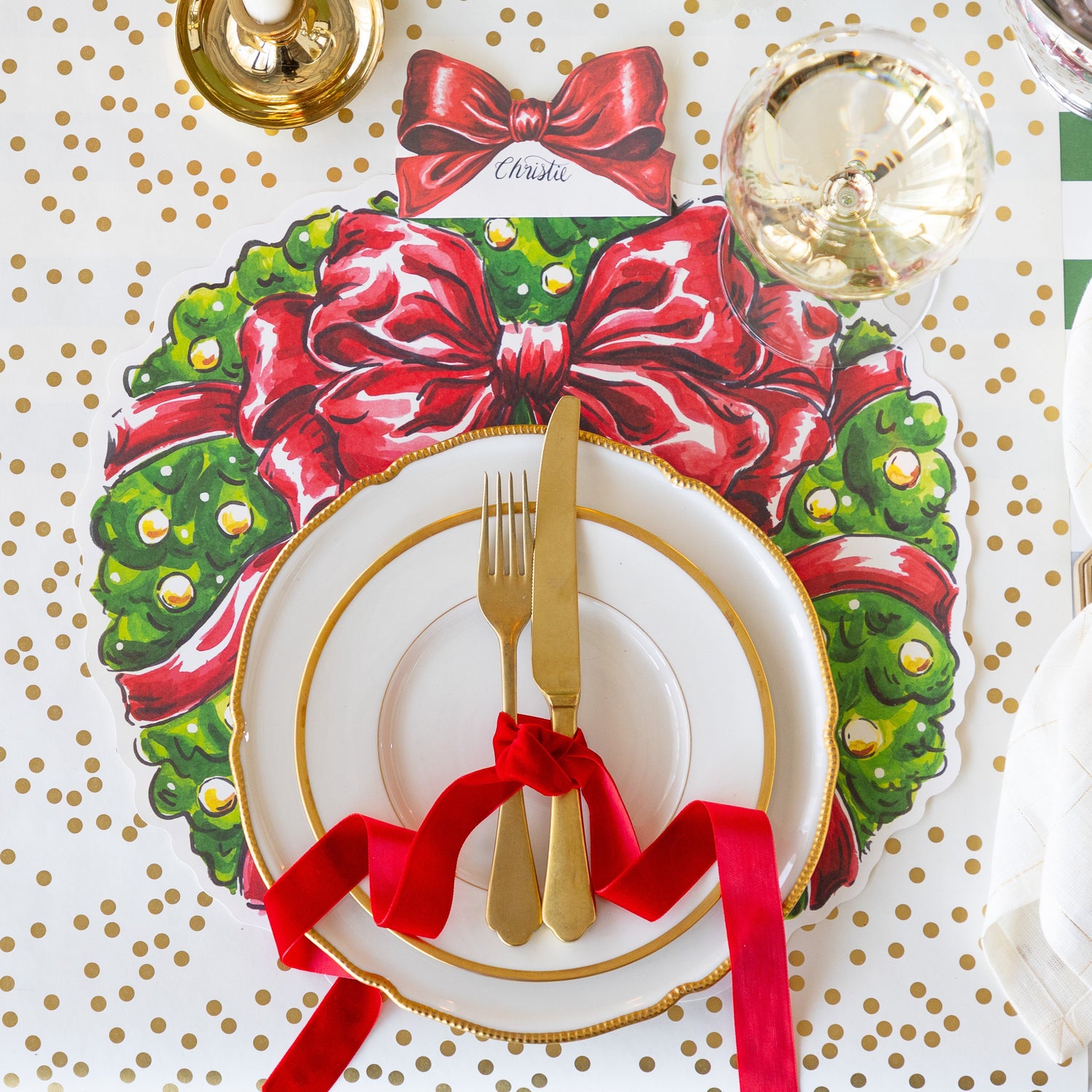 A place setting featuring the Die-cut Holiday Wreath Placemat with gold rimmed plates and a gold fork and knife tied in a red ribbon, atop, Bow Place Card reading &quot;Christie, and a glass of wine on the Gold Confetti Runner.
