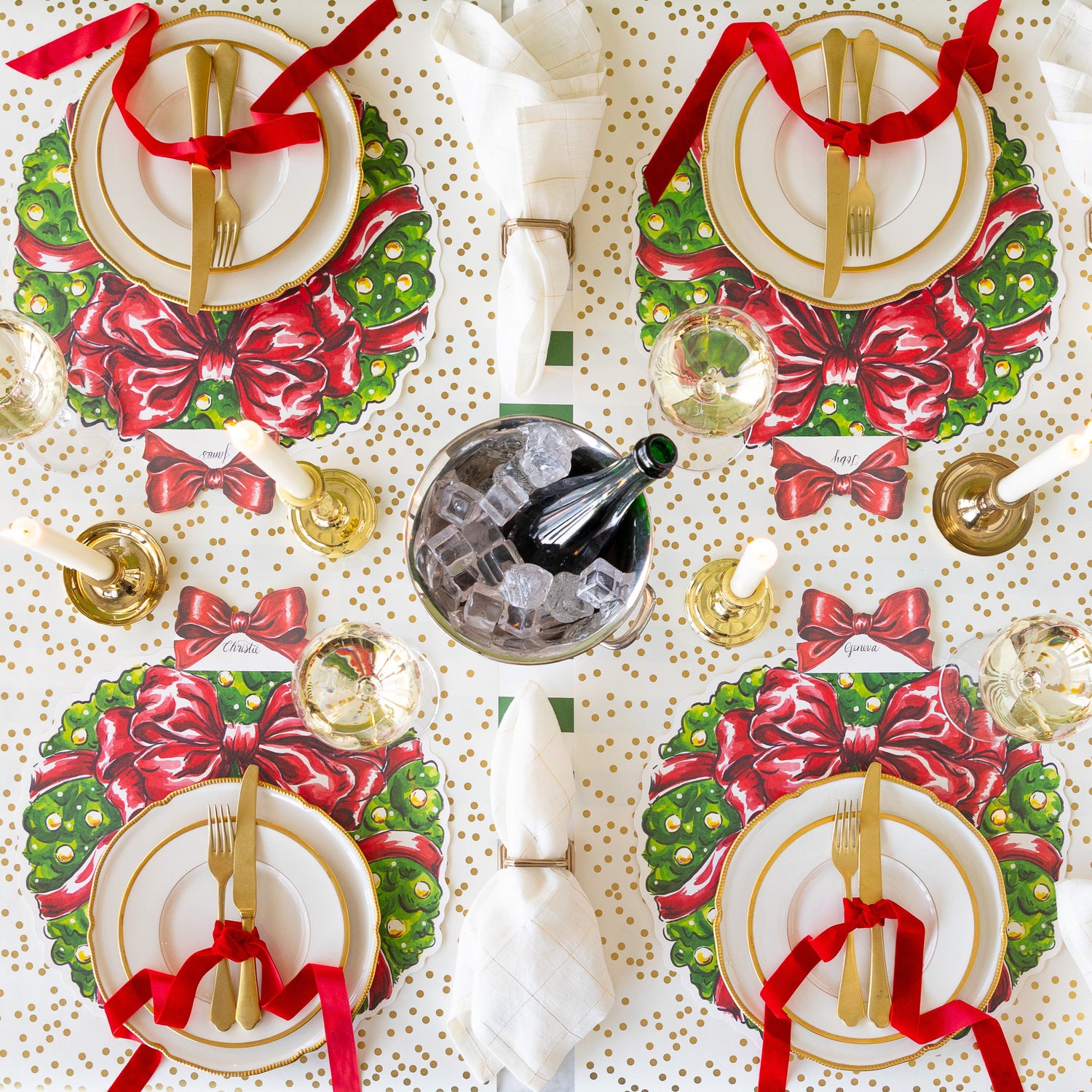 A table setting for four with lit candles and an ice bucket of champagne, featuring the Die-cut Holiday Wreath Placemats, gold rimmed dinnerware, gold flatware tied in a red velvet ribbon, and  napkins in the Napkin Ring with Place Card Holder on the Gold Confetti and Dark Green Classic Stripe Runner.
