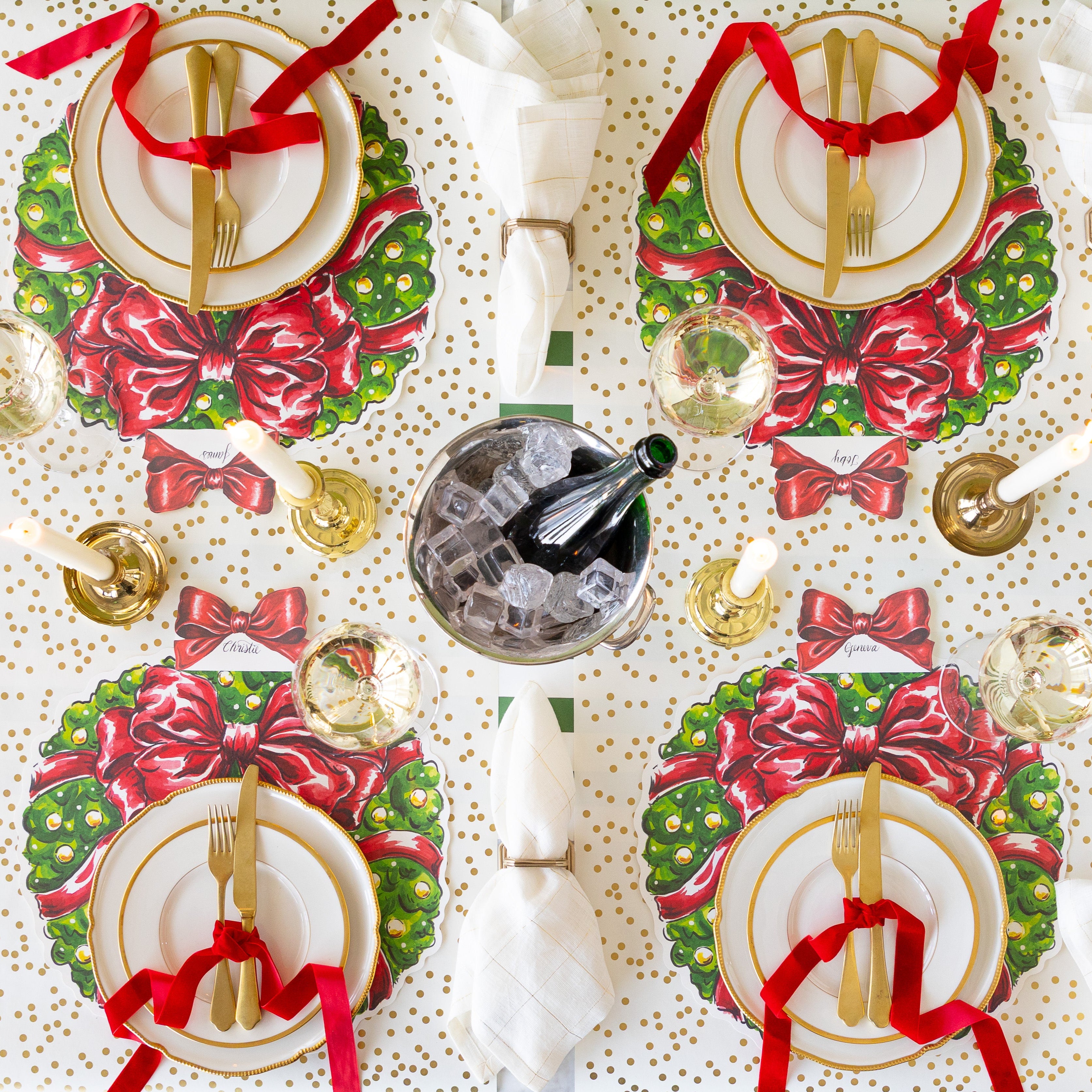 A table setting for four with lit candles and an ice bucket of champagne, featuring the Die-cut Holiday Wreath Placemats, gold rimmed dinnerware, gold flatware tied in a red velvet ribbon, and  napkins in the Napkin Ring with Place Card Holder on the Gold Confetti and Dark Green Classic Stripe Runner.