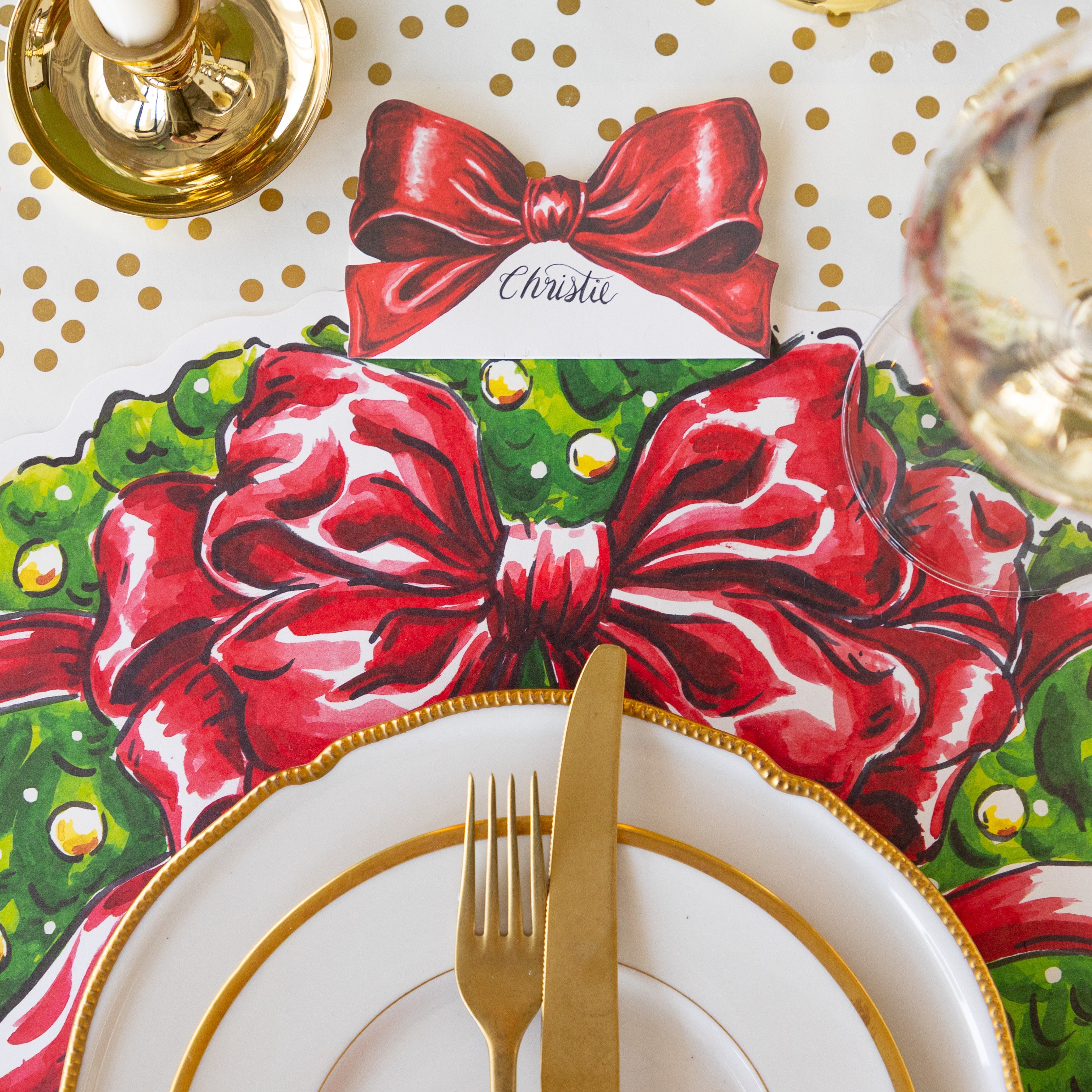 The Bow Place Card reading "Christie" behind a place setting with the Die-cut Holiday Wreath Placemat, gold rimmed dinner plates and a gold fork and knife on the Gold Confetti Runner.