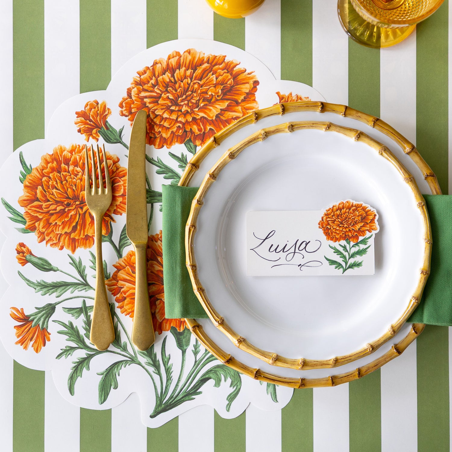 An elegant place setting featuring the Die-cut Marigold Bouquet Placemat offset to the left, under the Mepra Gold fork and knife, a green napkin in between Juliska Bamboo Dinner Plates with the Marigold Bloom Place Card on top, Classic Moss Stripe Runner vertically and Rattan Weave horizontally underneath the setting.