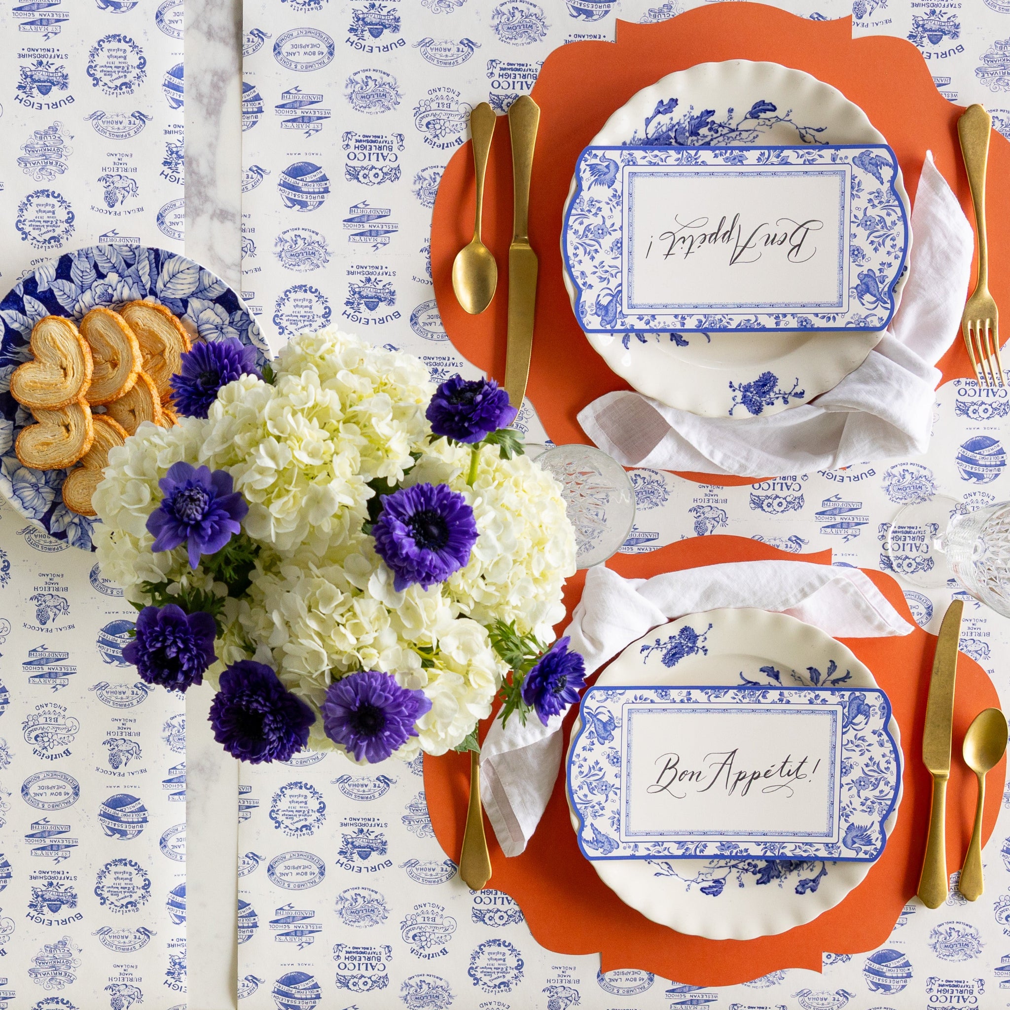 A table set for two with blue and white plates on Die-cut Orange French Frame Placemats gold cutlery, a bouquet of white and purple flowers, and plate of cookies on the Burleigh Heritage Runner. The plates feature Blue Regal Peacock Table Accents reading &quot;Bon Appétit!&quot; atop.