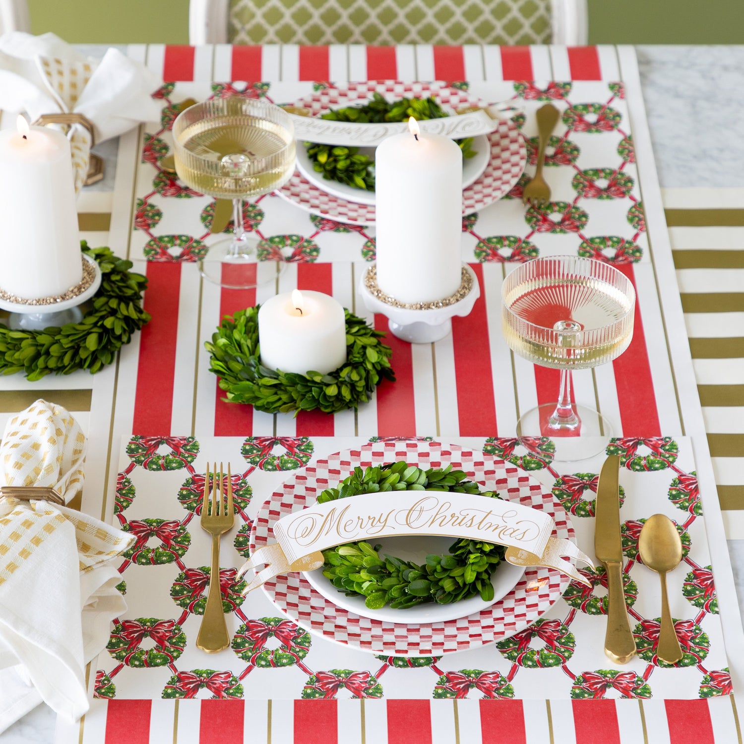 A table setting with lit candles and boxwood wreaths around them, Holiday Wreath Lattice Placemats, red-and-white checkered plates with boxwood wreaths and Merry Christmas Banner Table Accents atop, gold flatware, and champagne coupe glasses on the Red & Gold Awning Stripe and Gold Classic Stripe Runners.