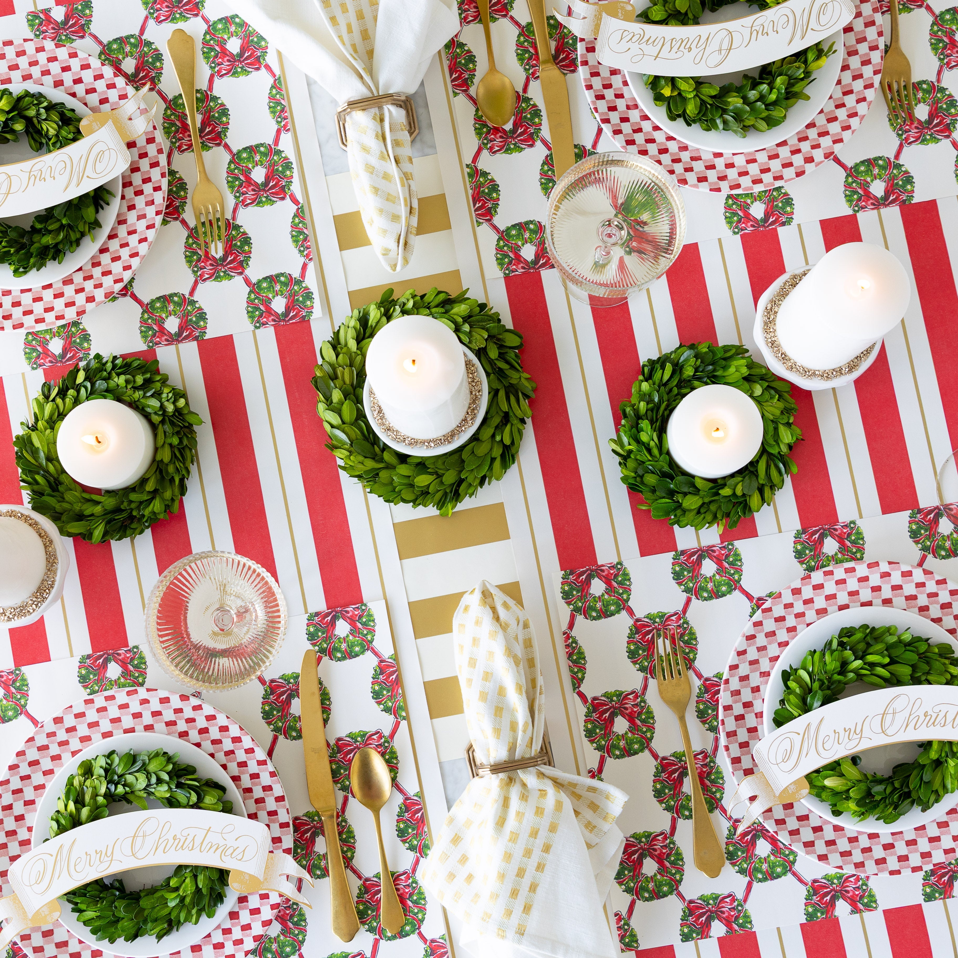 An elegant Christmas table setting with candles, boxwood wreath, Holiday Wreath Lattice Placemats, Merry Christmas Banner Table Accents, and napkins in a holder on the Red & Gold Awning Stripe and Gold Classic Stripe Runners.