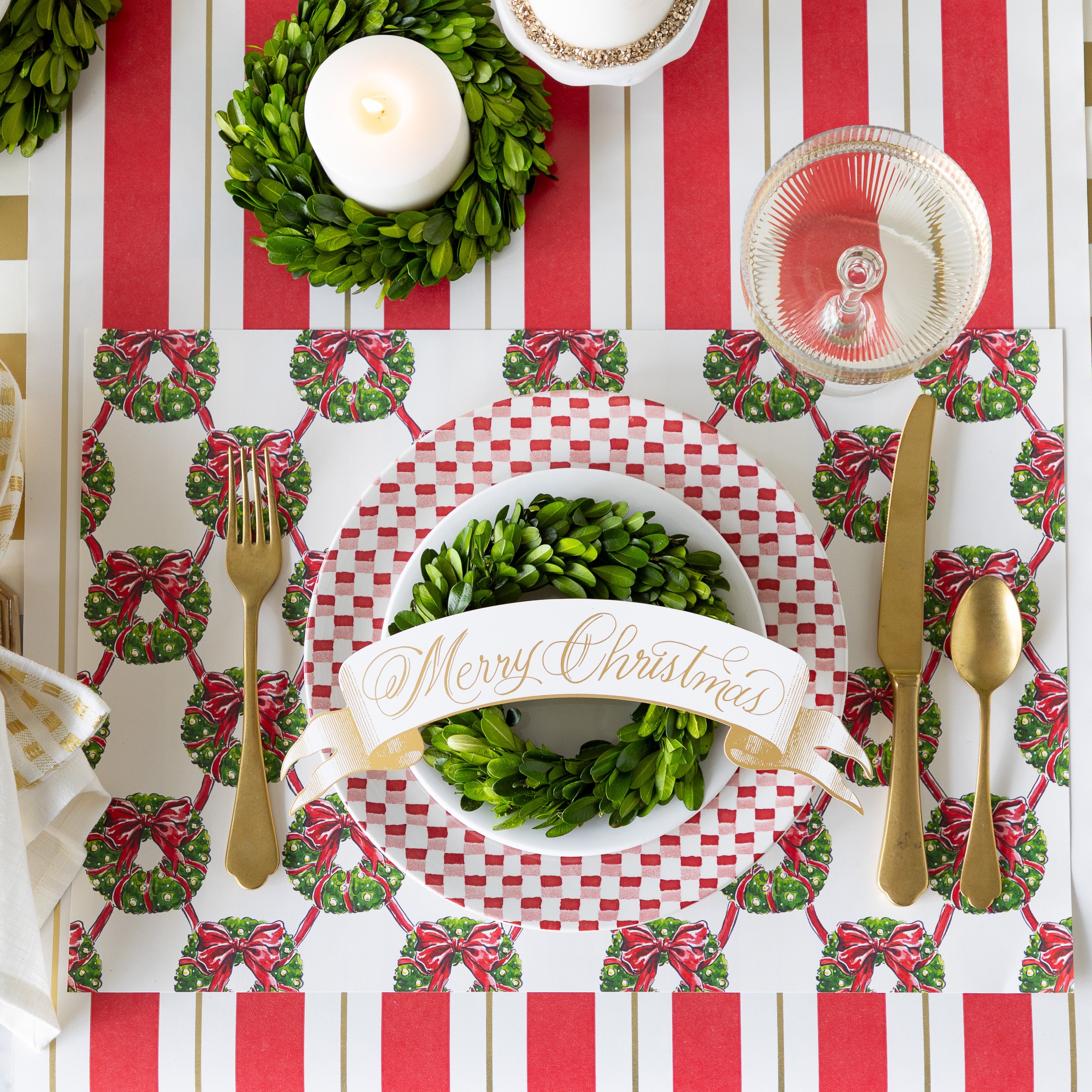 A place setting featuring the Holiday Wreath Lattice Placemat, a red-and-white checkered plate with a white plate, boxwood wreath and Merry Christmas Banner Table Accent atop, gold flatware, and coupe glass on the Red & Gold Awning Stripe Runner.