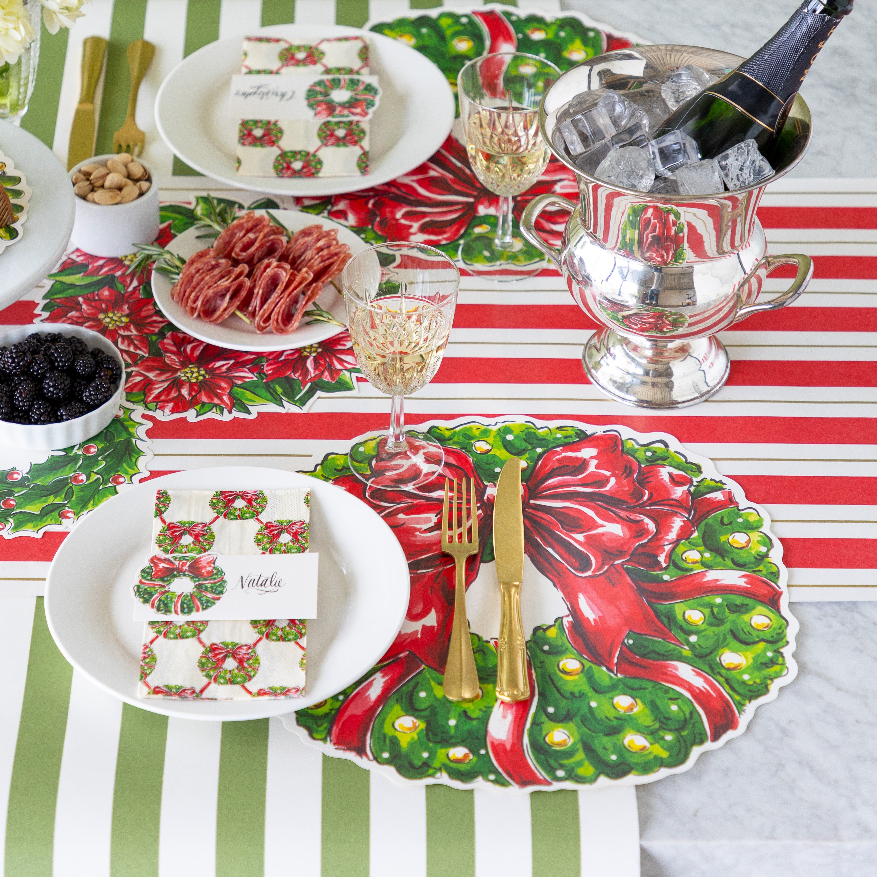 A festive table setting with charcuterie and champagne on ice, featuring the Die-cut Holiday Wreath Placemat, a plate with the Holiday Wreath Guest Napkin and Holiday Wreath Place Card atop, and gold cutlery on the Red & Gold Awning Stripe Runner and Moss Classic Stripe Runner.