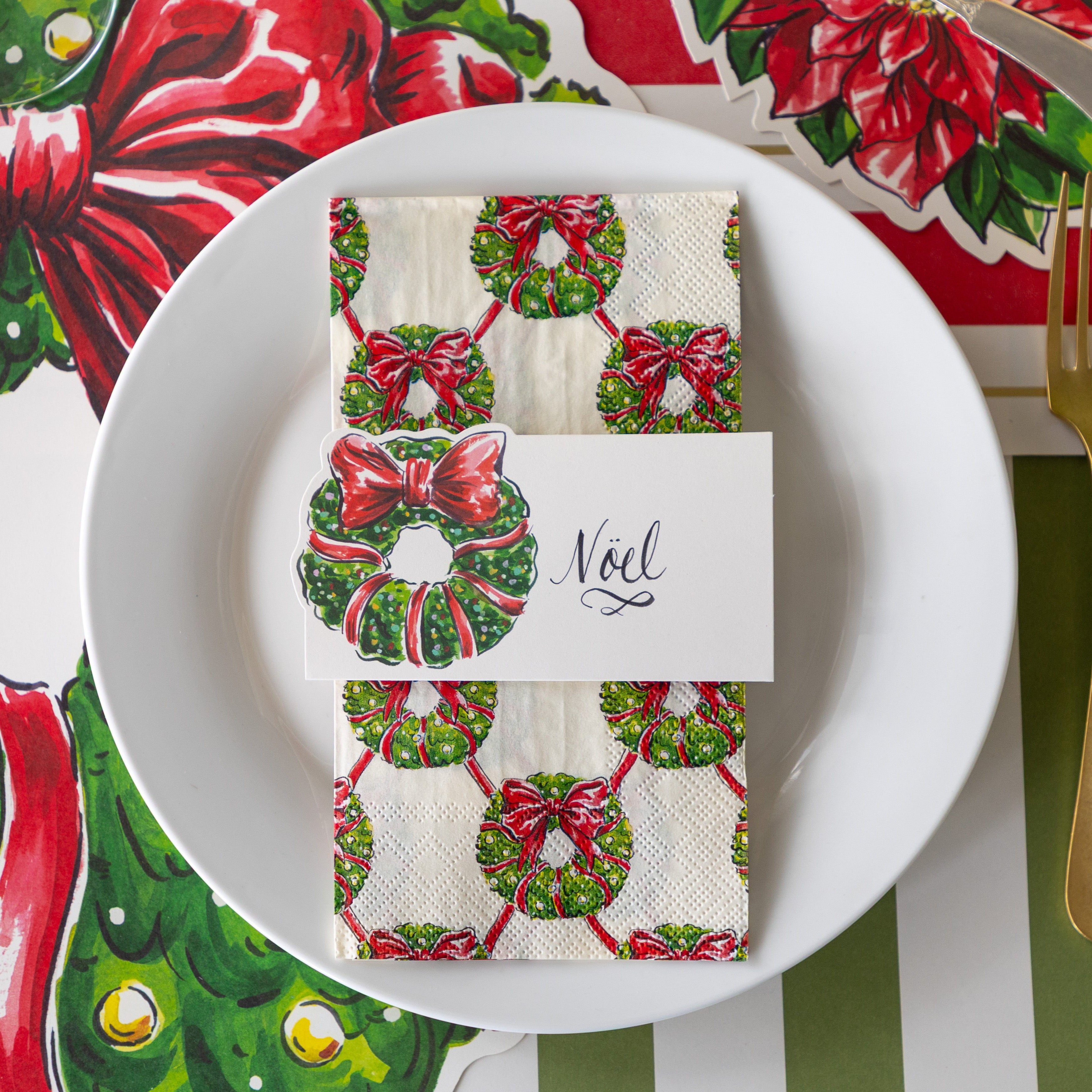 A place setting with the Holiday Wreath Place Card reading &quot;Noel&quot;, atop the Holiday Wreath Guest Napkin and white plate, on the Die-cut Holiday Wreath Placemat.