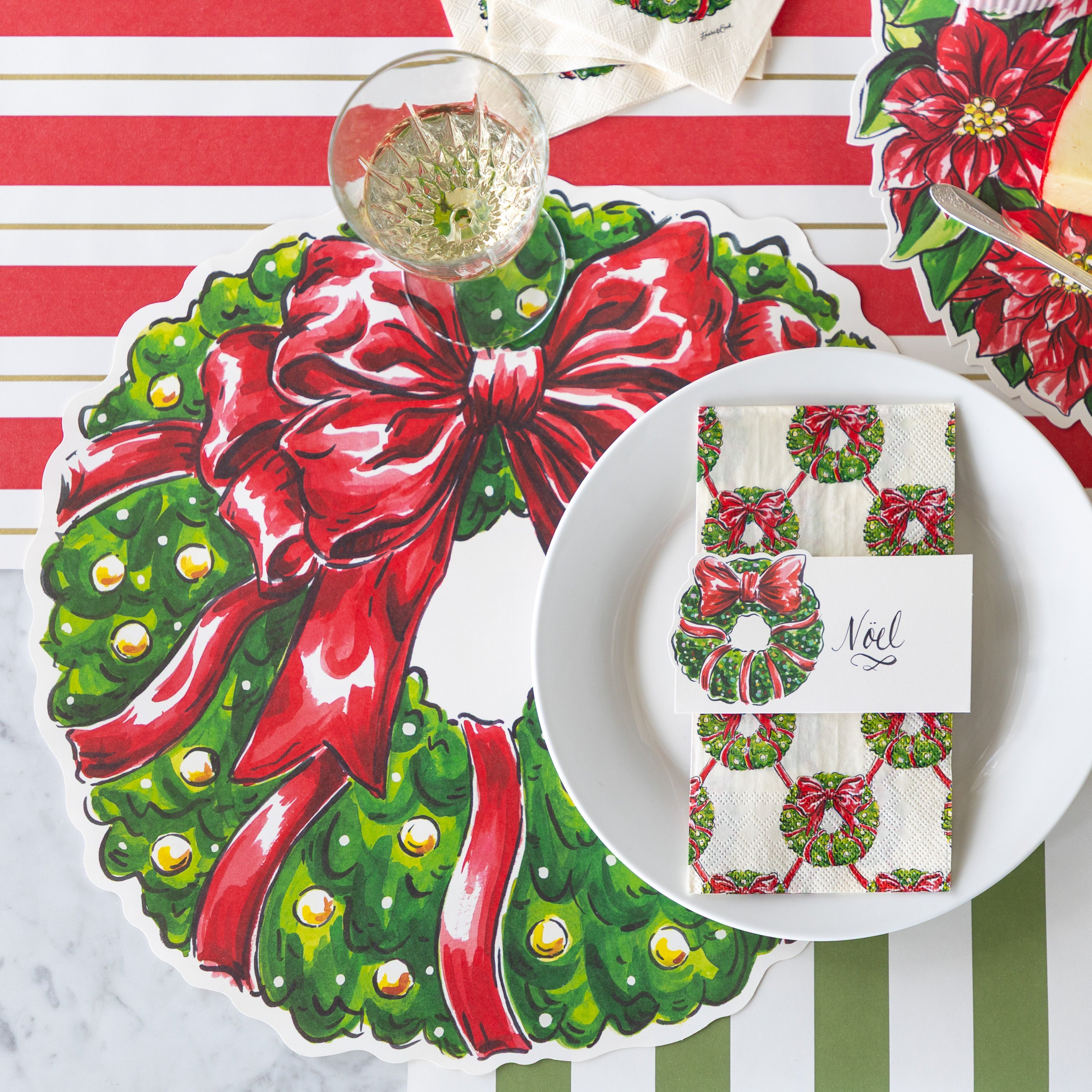 A place setting featuring the Die-cut Holiday Wreath Placemat, a plate with the Holiday Wreath Guest Napkin and Wreath Place Card reading "Noel" atop, Moss Classic Stripe and Red & Gold Awning Stripe Runner underneath.
