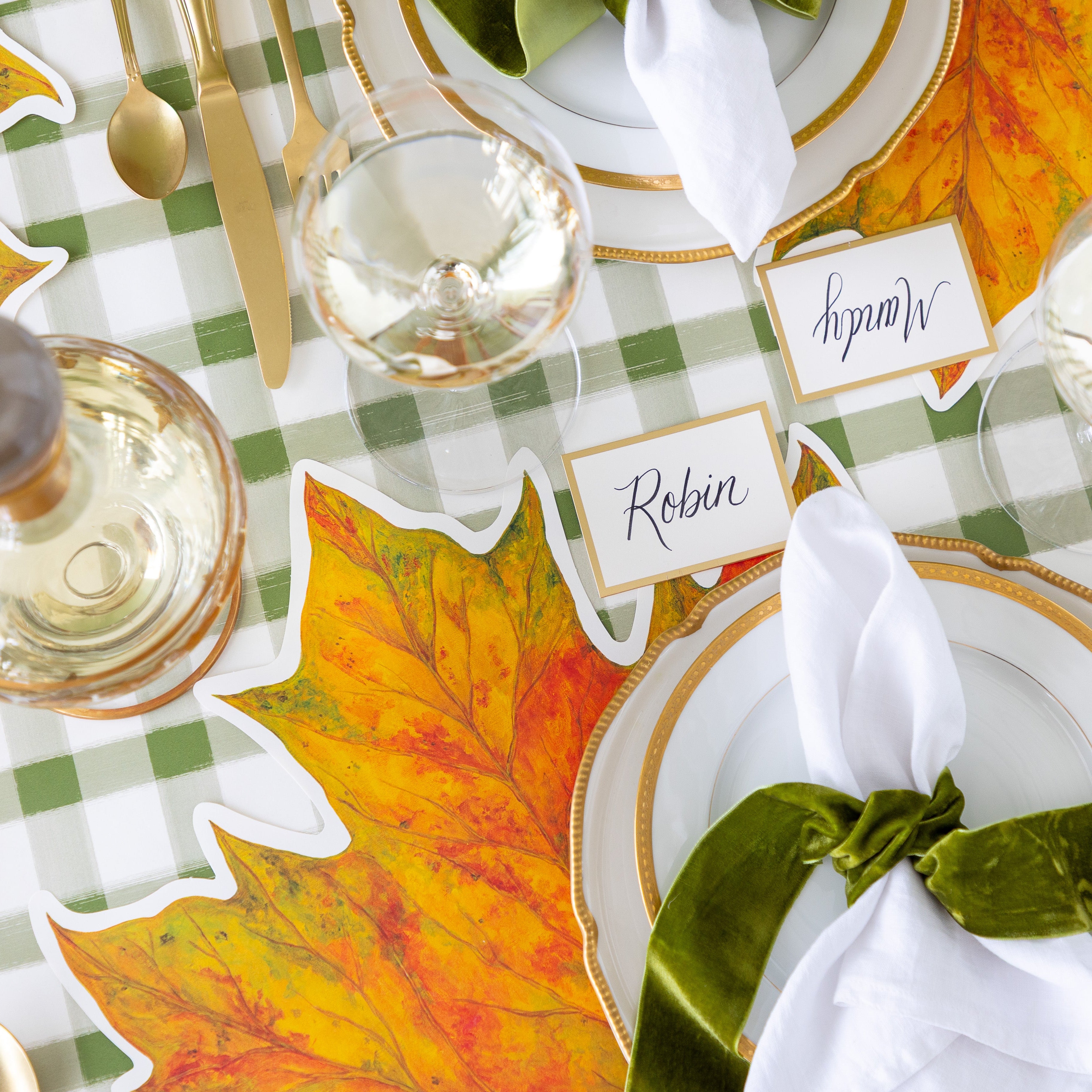 An elegant place setting featuring the Die-cut Fall Leaf Placemat, gold rimmed plates, the Gold Frame Place Card reading "Robin", white napkins tied by a green velvet ribbon and the Green Painted Check Runner underneath.