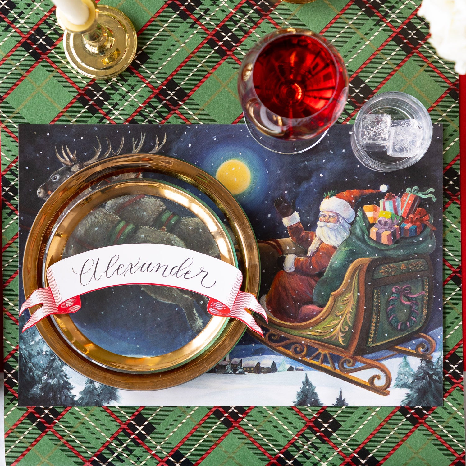 A Christmas place setting featuring the Sleigh Ride Placemat, gold rimmed dinner plates with the Classic Red Banner Table Accent reading &quot;Alexander&quot; atop, a glass of wine, a candle and Green Plaid Runner underneath.