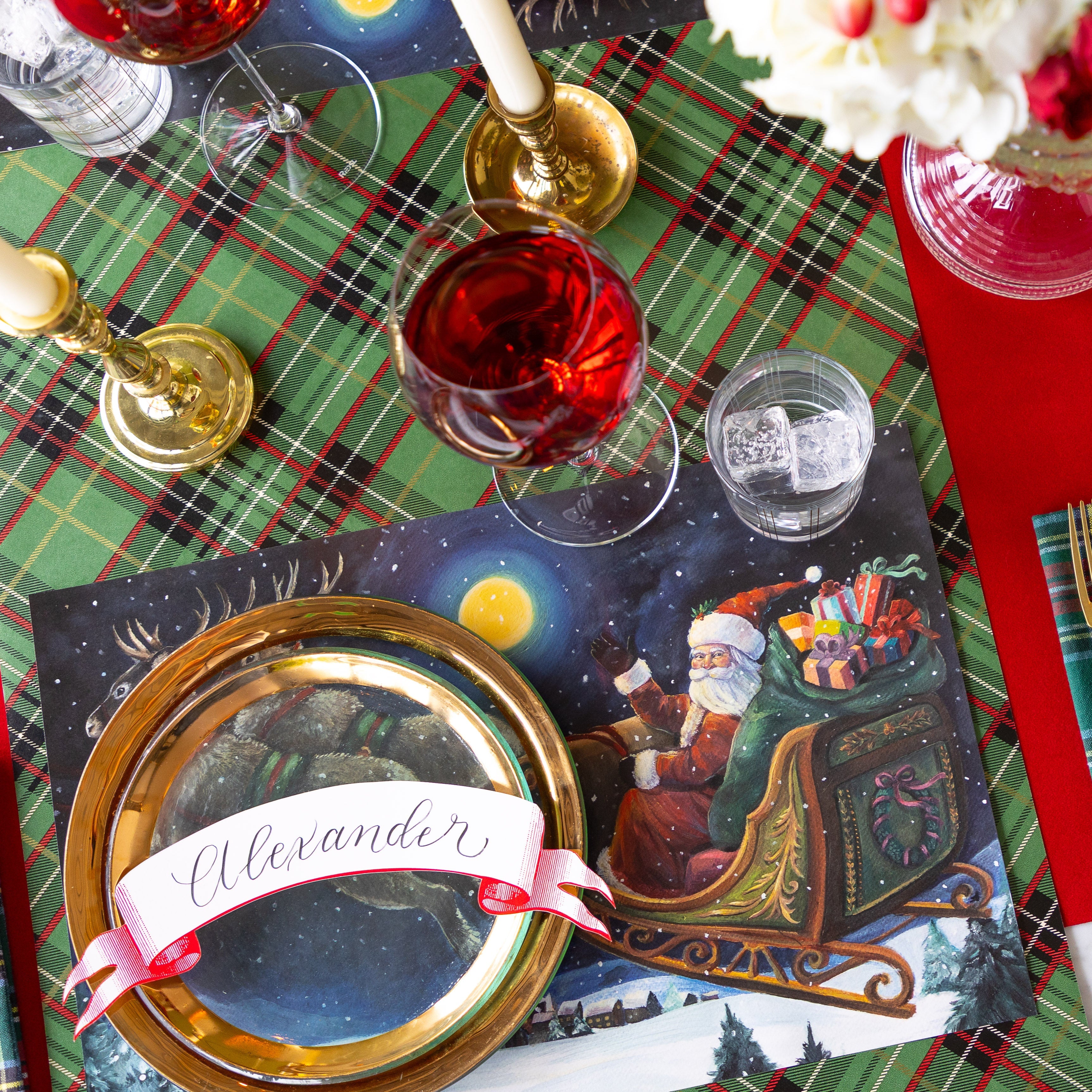 A Christmas place setting featuring the Sleigh Ride Placemat, gold rimmed dinner plates with the Classic Red Banner Table Accent reading &quot;Alexander&quot; atop, a glass of wine, candles and Green Plaid Runner underneath.