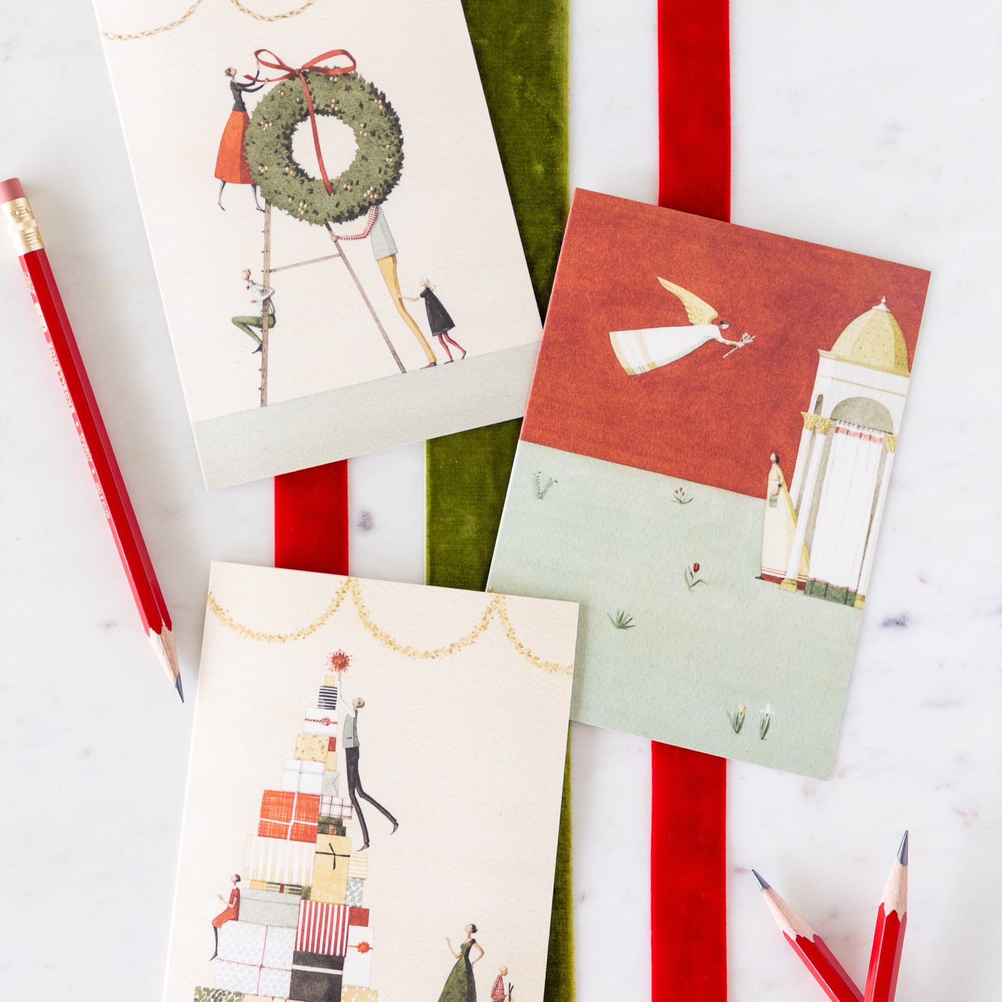 Laura Stoddart holiday greeting cards arranged on a table on top of red and green velvet ribbons and pencils nearby.