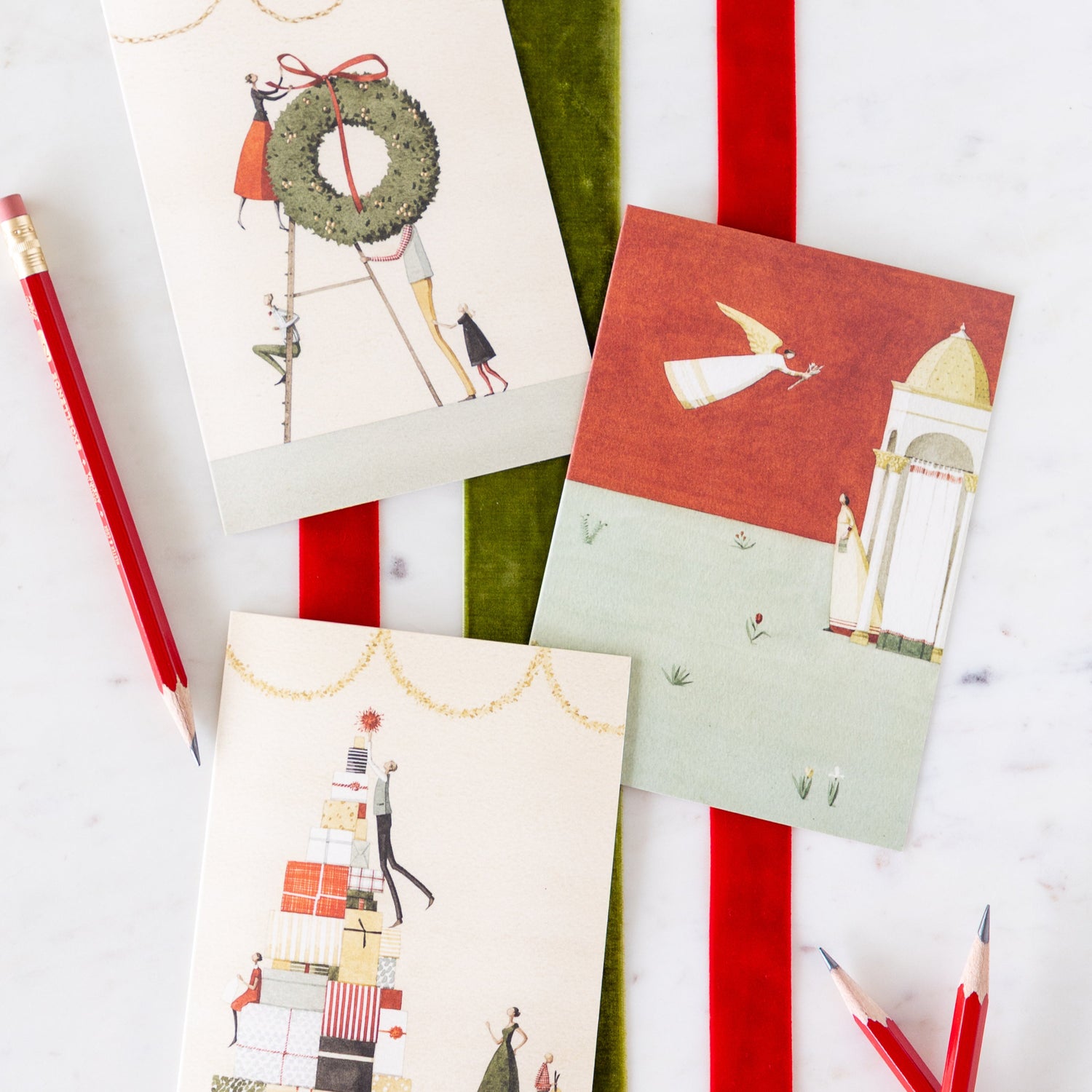 Laura Stoddart holiday greeting cards arranged on a table on top of red and green velvet ribbons and pencils nearby.