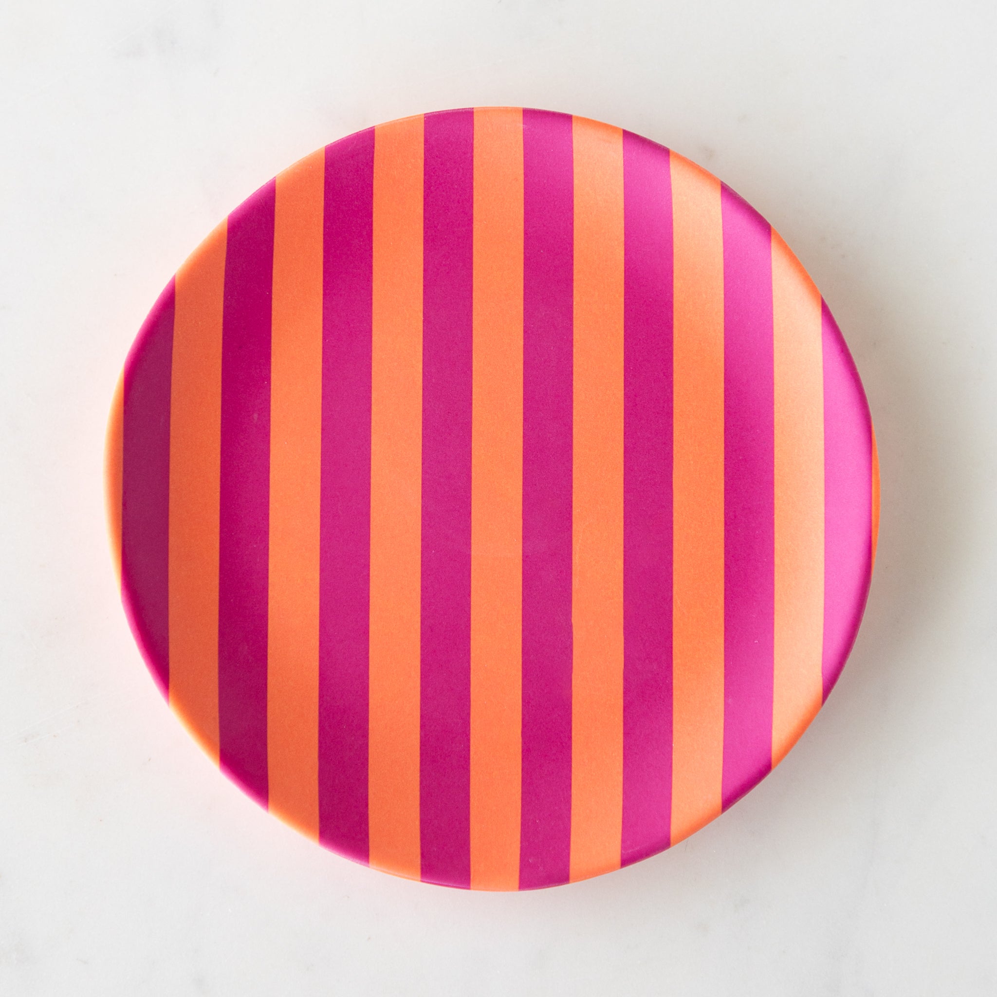 A round Stripe Melamine Plate with alternating vertical orange and magenta stripes on a marble table.