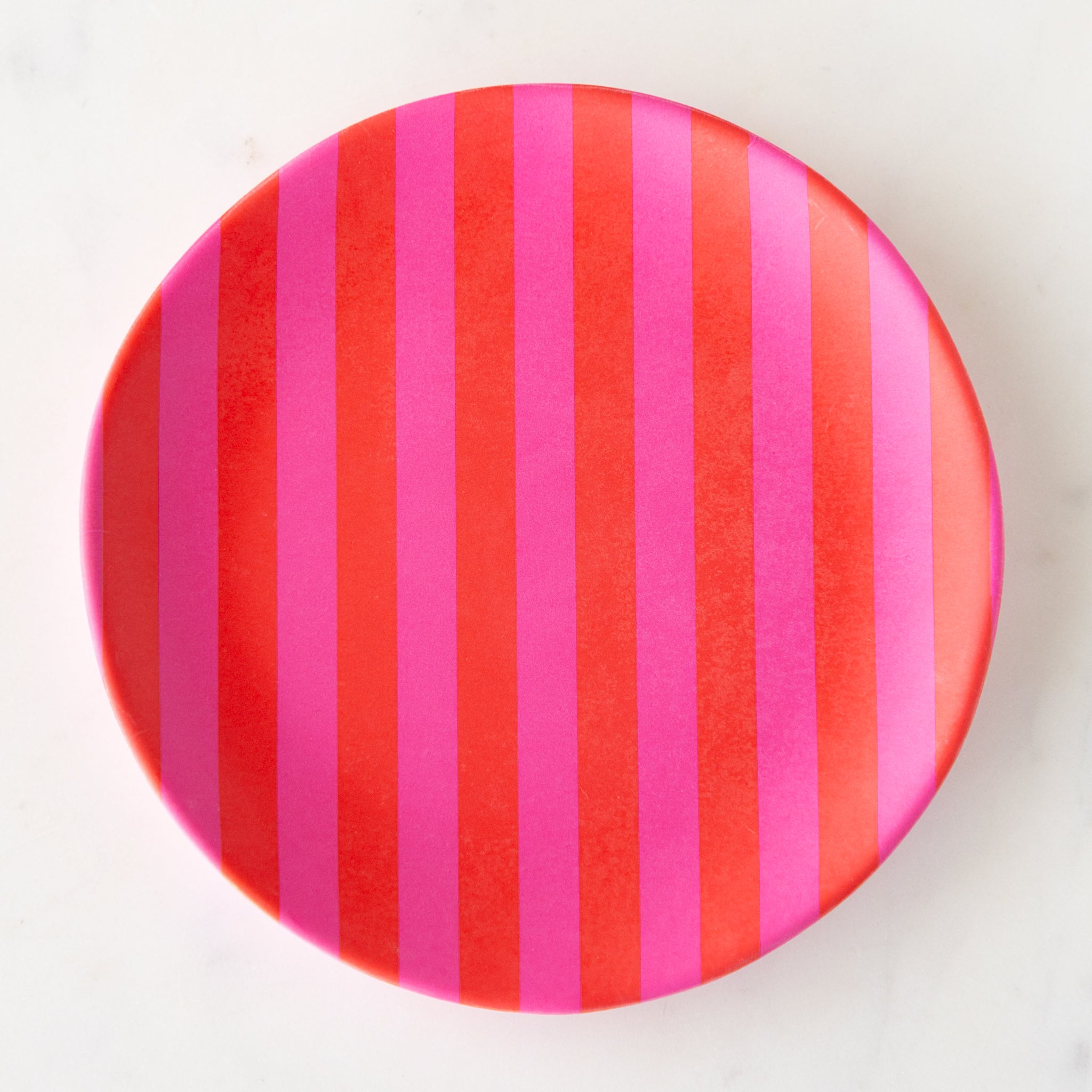 A round Stripe Melamine Plate with alternating vertical hot pink and red stripes on a marble table.