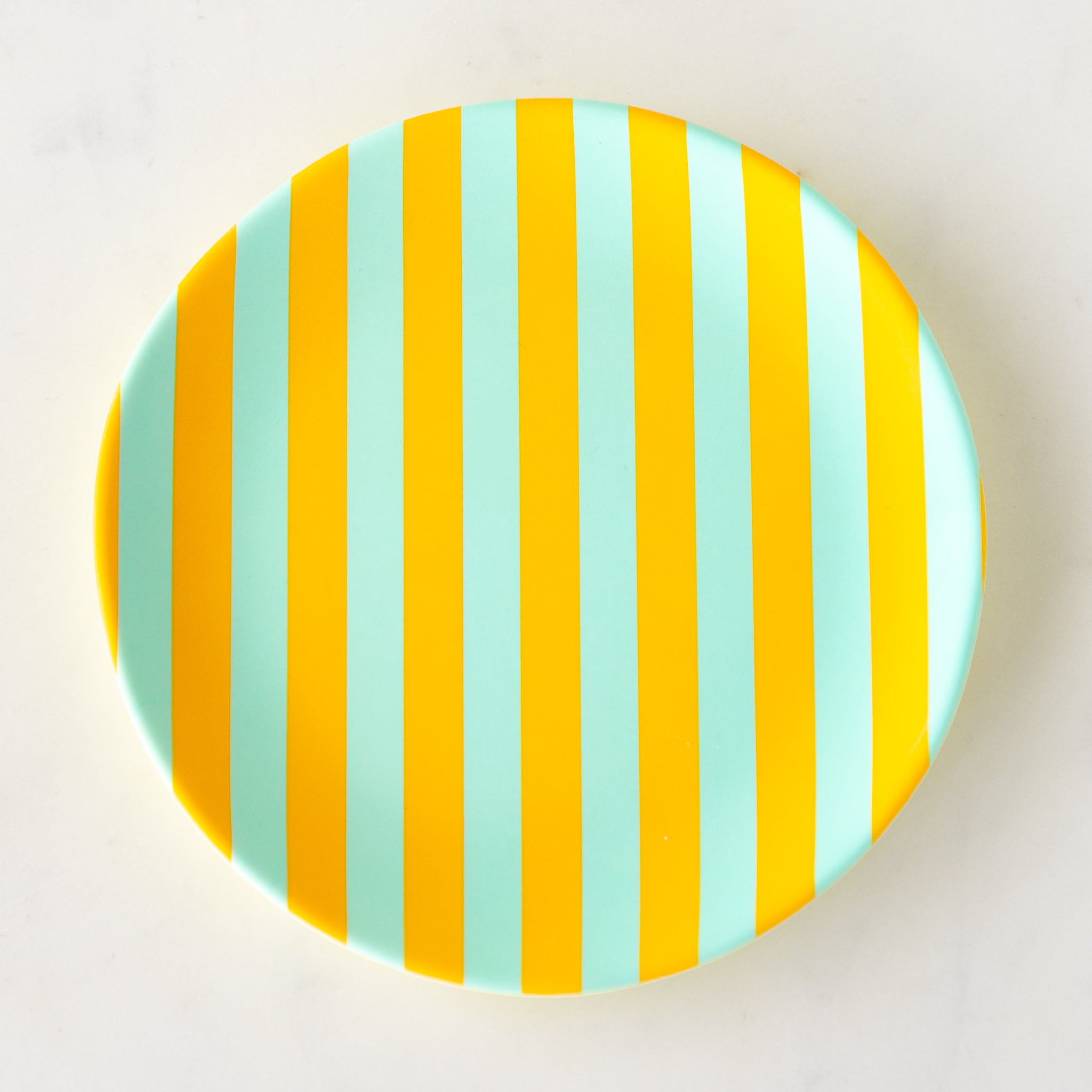 A round Stripe Melamine Plate with alternating vertical marigold and seafoam stripes on a marble table.