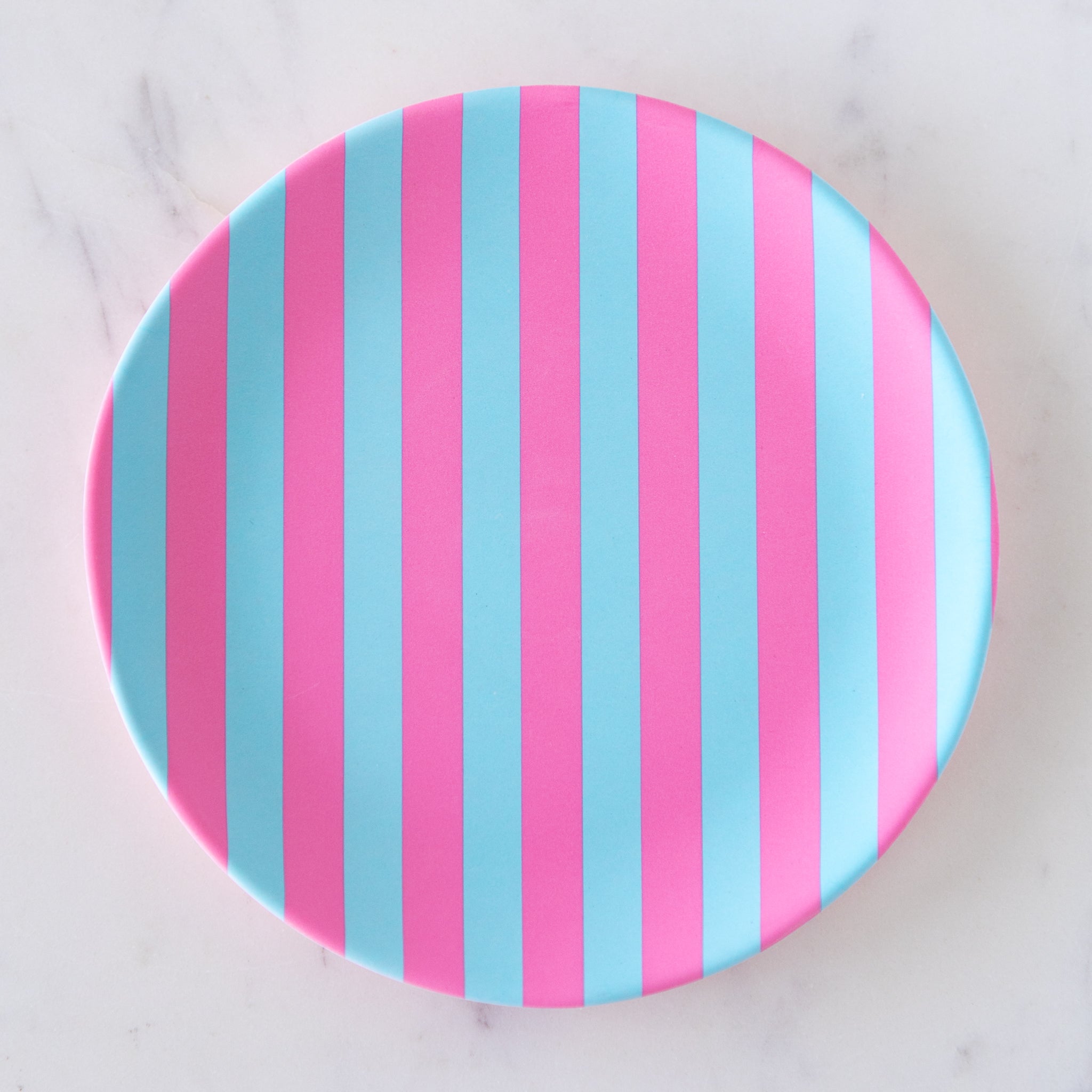 A round Stripe Melamine Plate with alternating vertical pink and light blue stripes on a marble table.