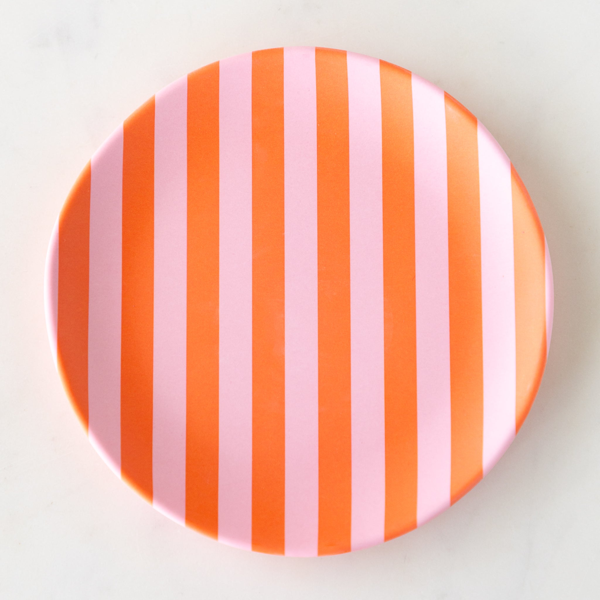A round Stripe Melamine Plate with alternating vertical pink and orange stripes on a marble table.
