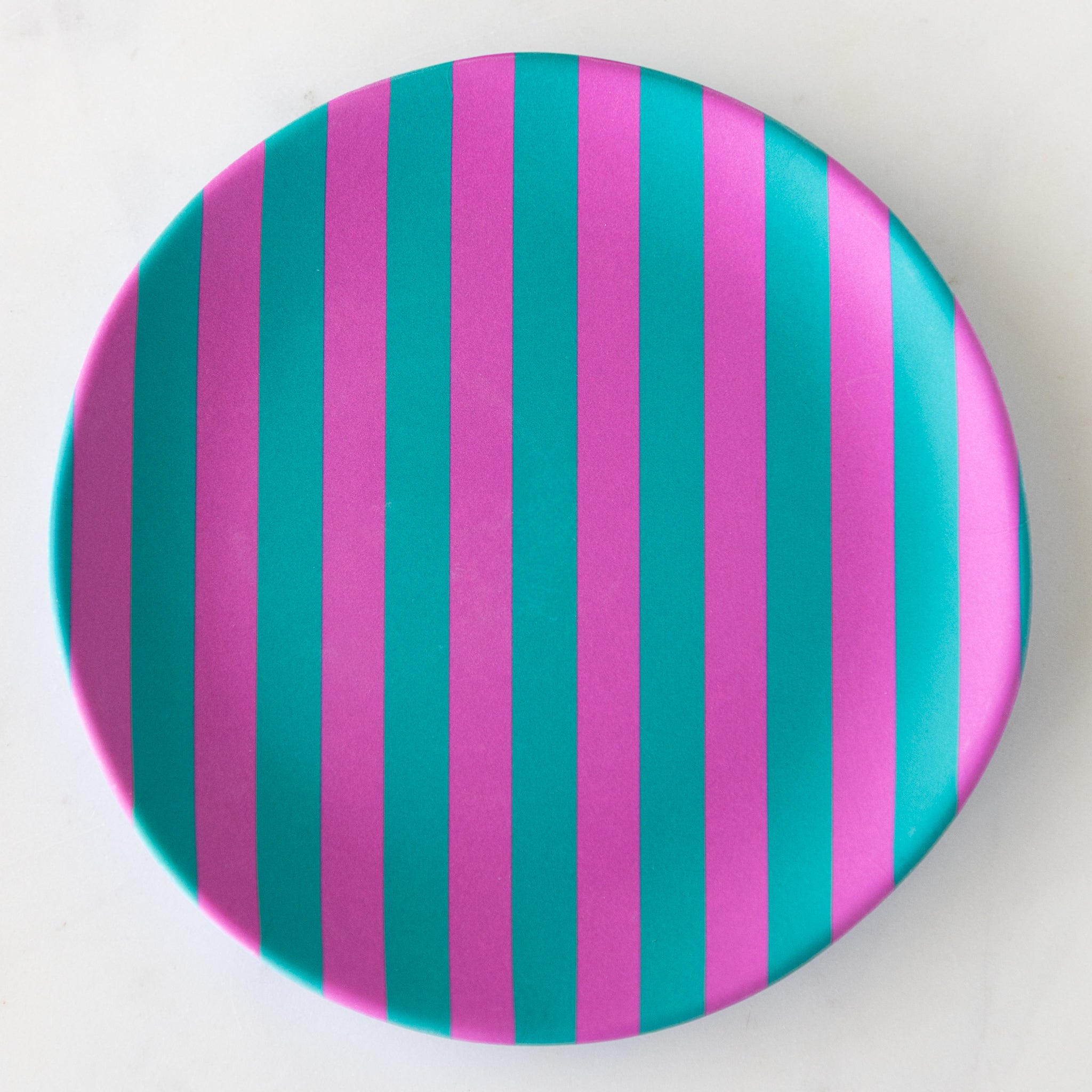 A round Stripe Melamine Plate with alternating vertical fuchsia and turquoise stripes on a marble table.
