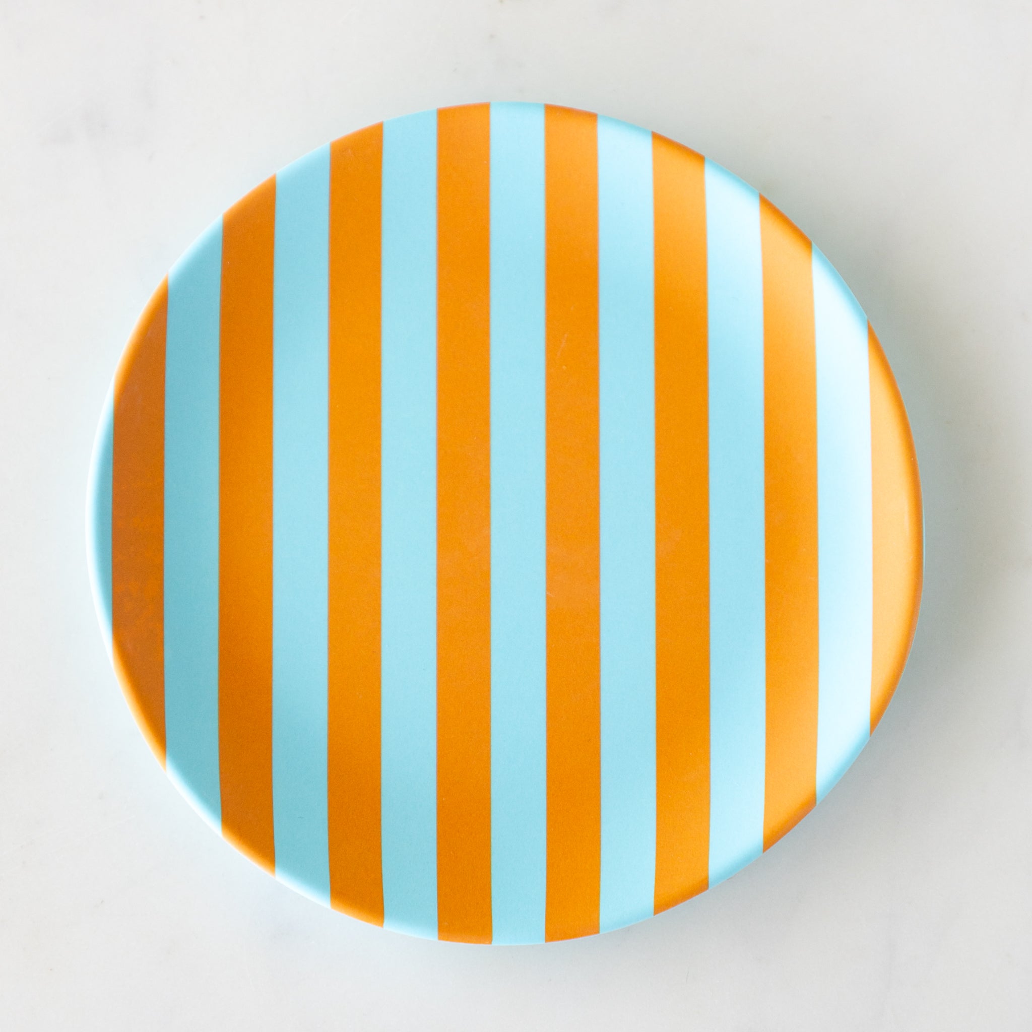 A round Stripe Melamine Plate with alternating vertical light blue and orange stripes on a marble table.