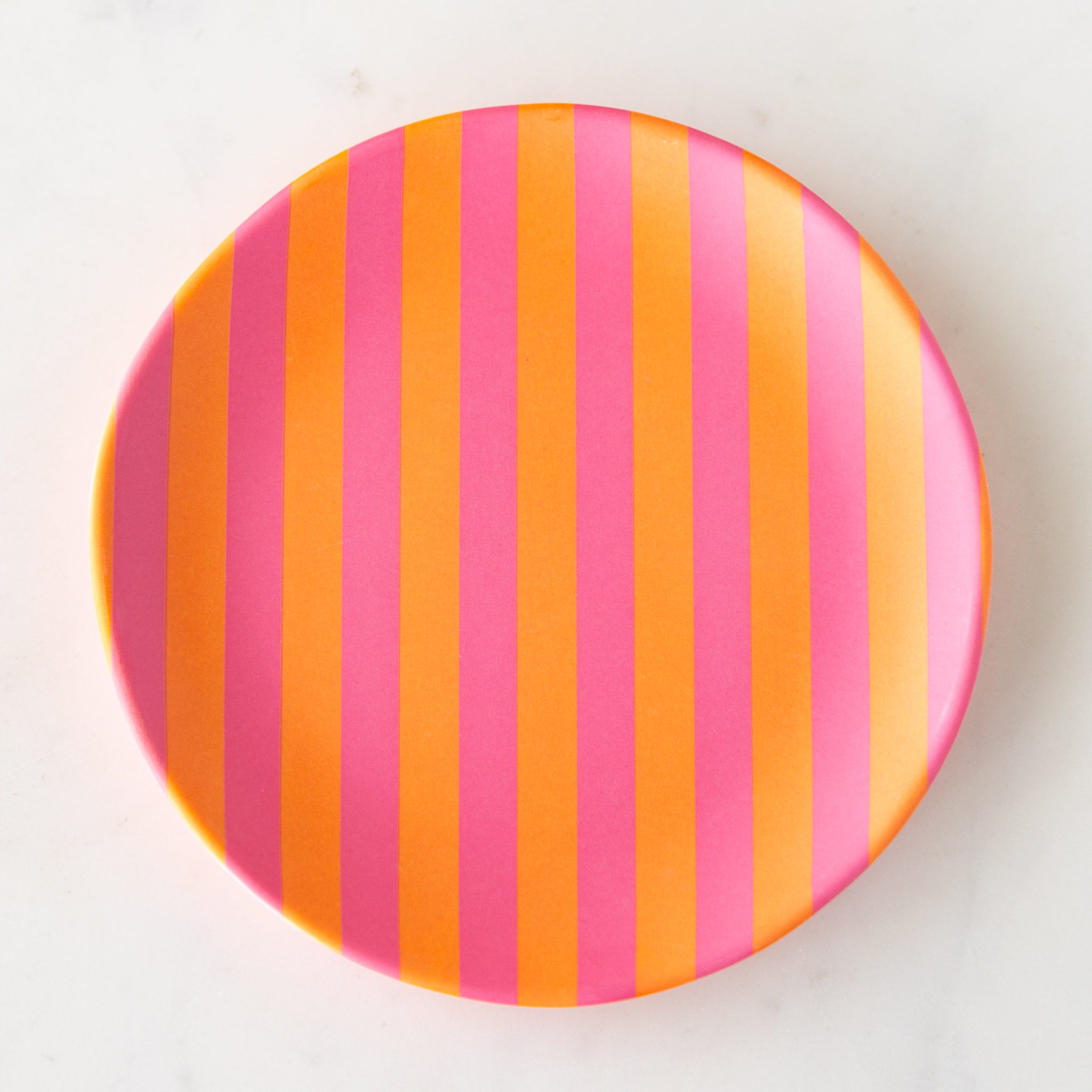 A round Stripe Melamine Plate with alternating vertical pink and orange stripes on a marble table.
