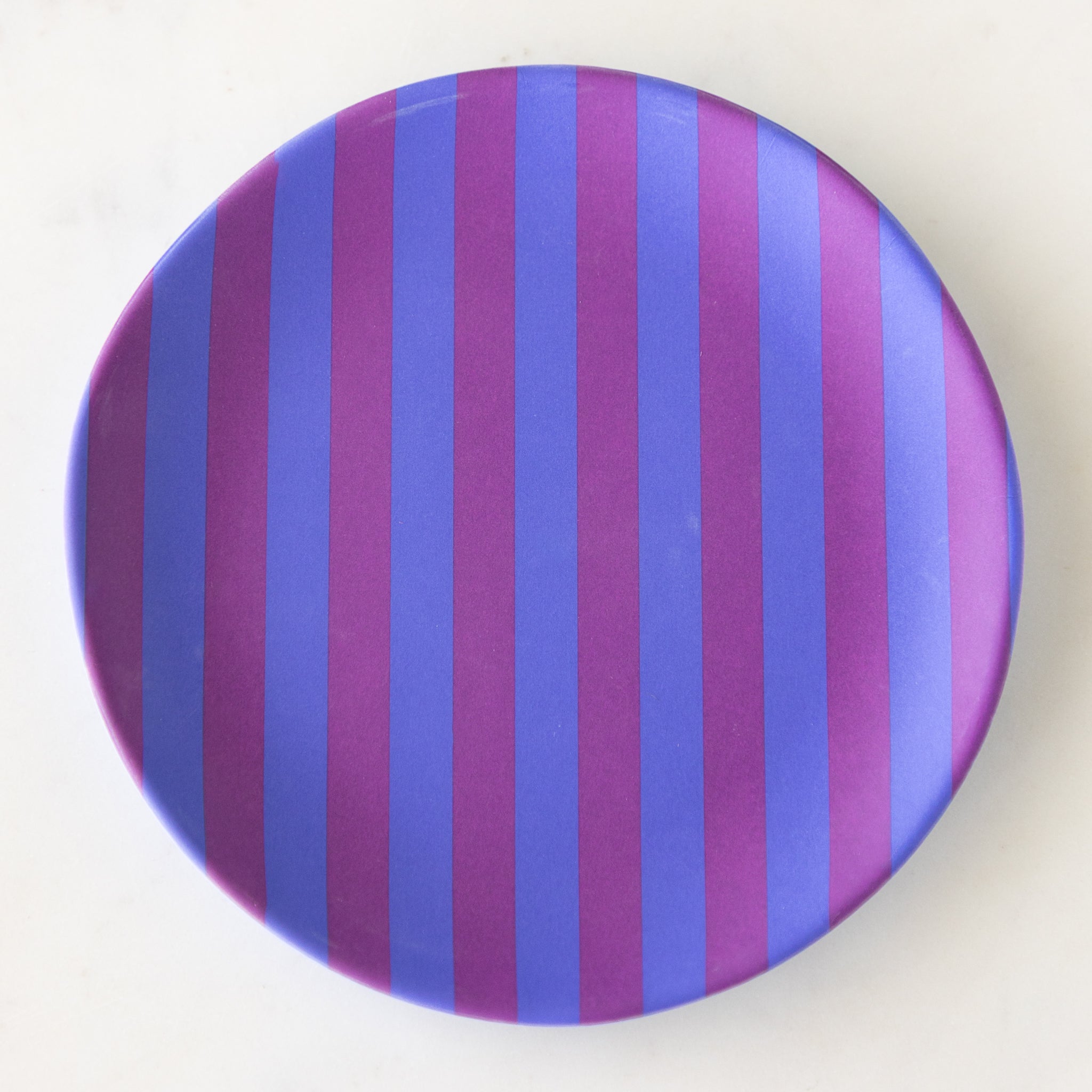 A round Stripe Melamine Plate with alternating vertical purple and blue stripes on a marble table.