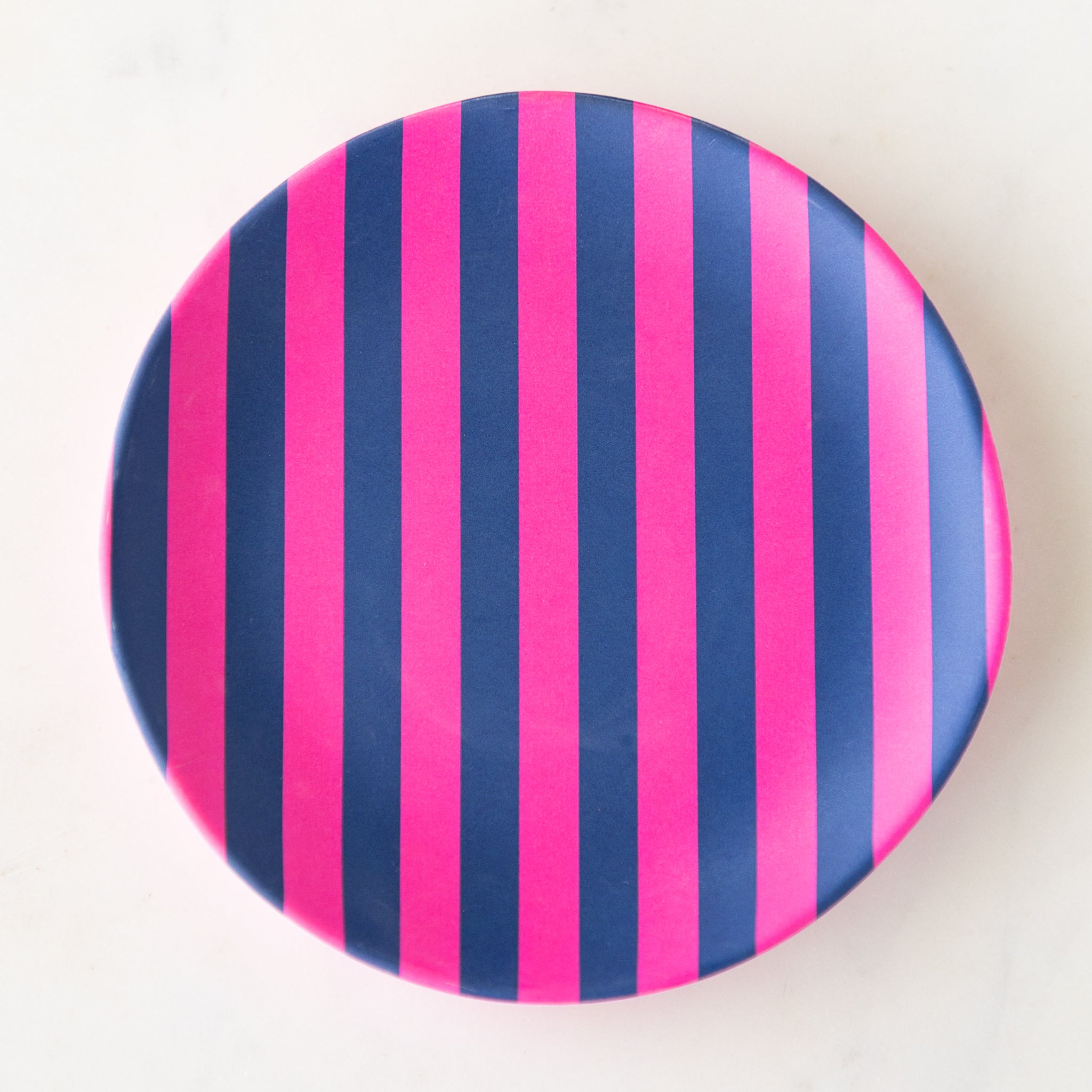 A round Stripe Melamine Plate with alternating vertical hot pink and navy stripes on a marble table.