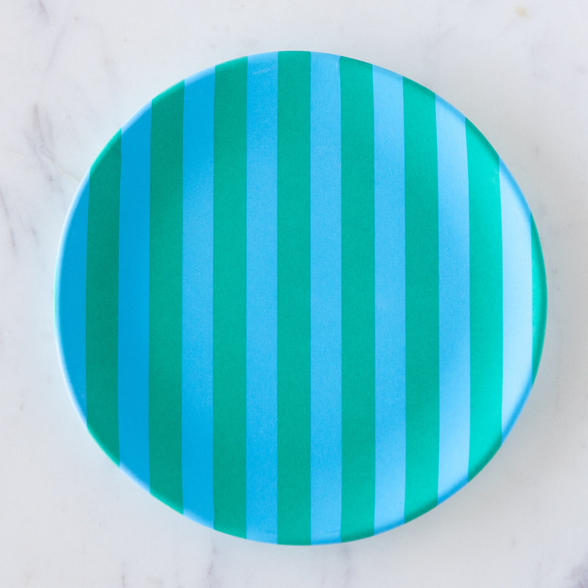 A round Stripe Melamine Plate with alternating vertical light blue and turquoise stripes on a marble table.