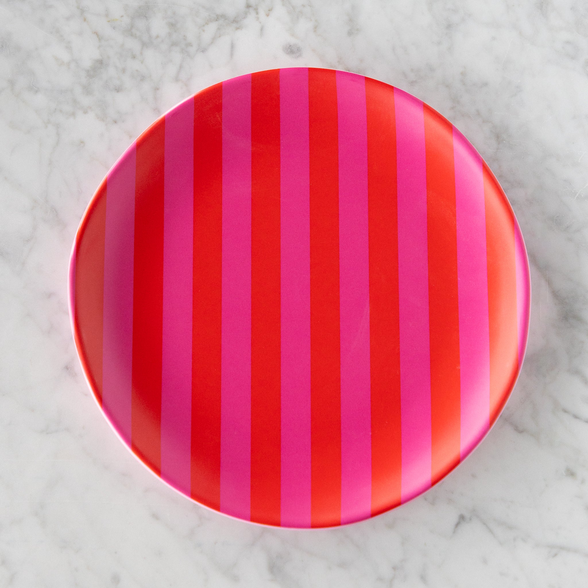 A round Stripe Melamine Plate with alternating vertical hot pink and red stripes on a marble table.