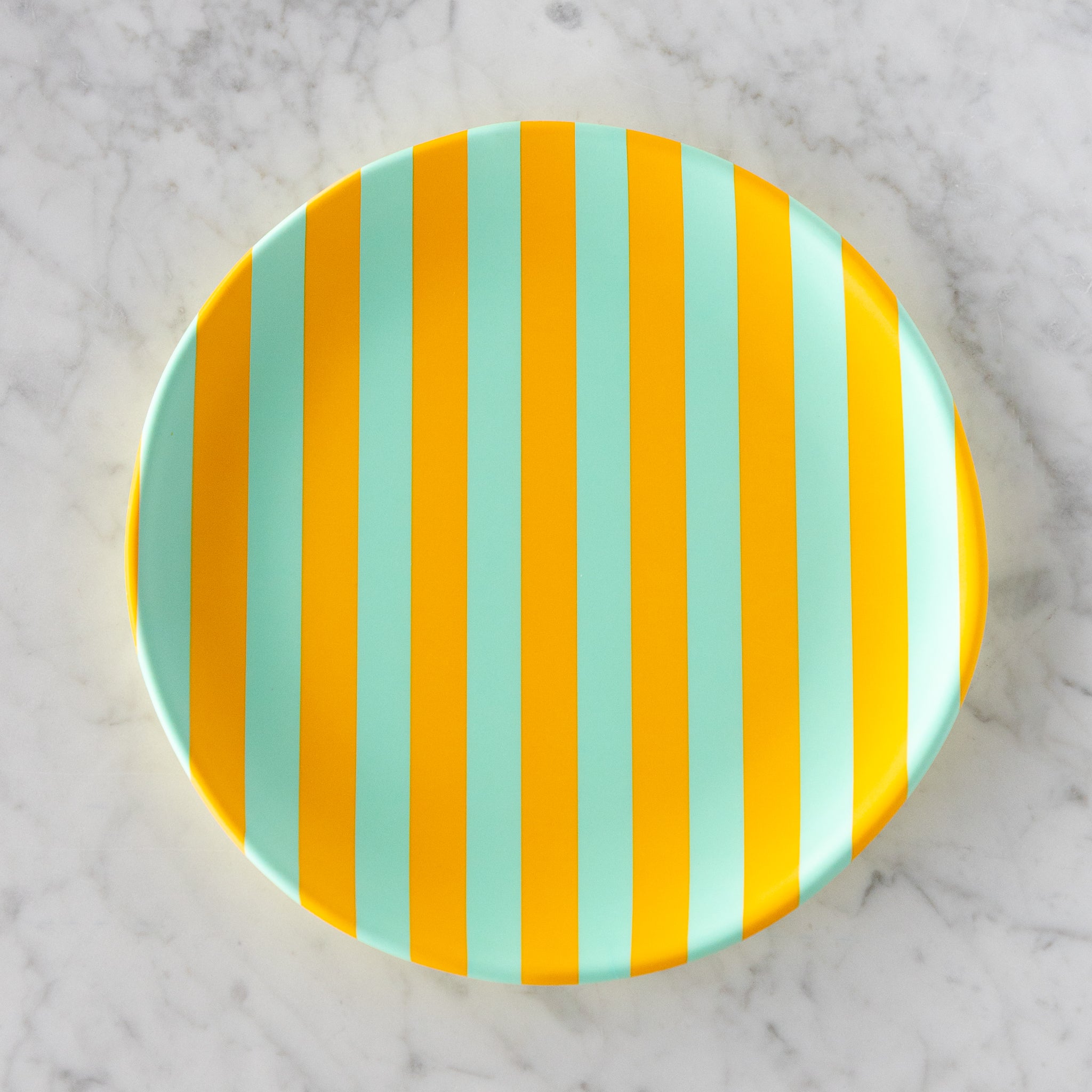 A round Stripe Melamine Plate with alternating vertical marigold and seafoam stripes on a marble table.