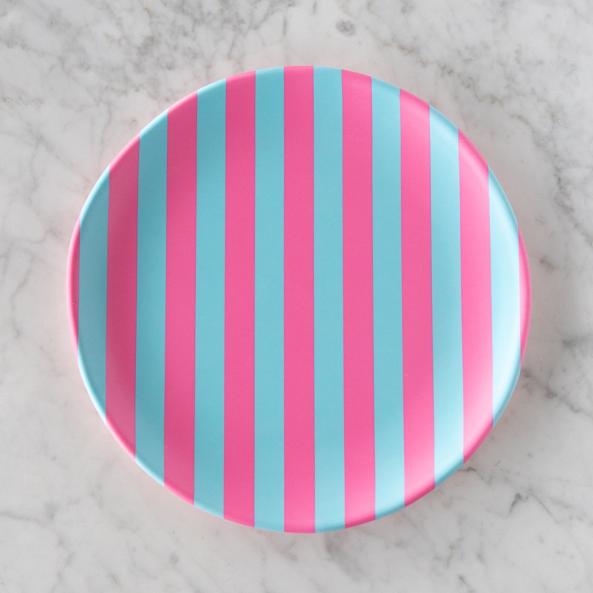 A round Stripe Melamine Plate with alternating vertical pink and light blue stripes on a marble table.