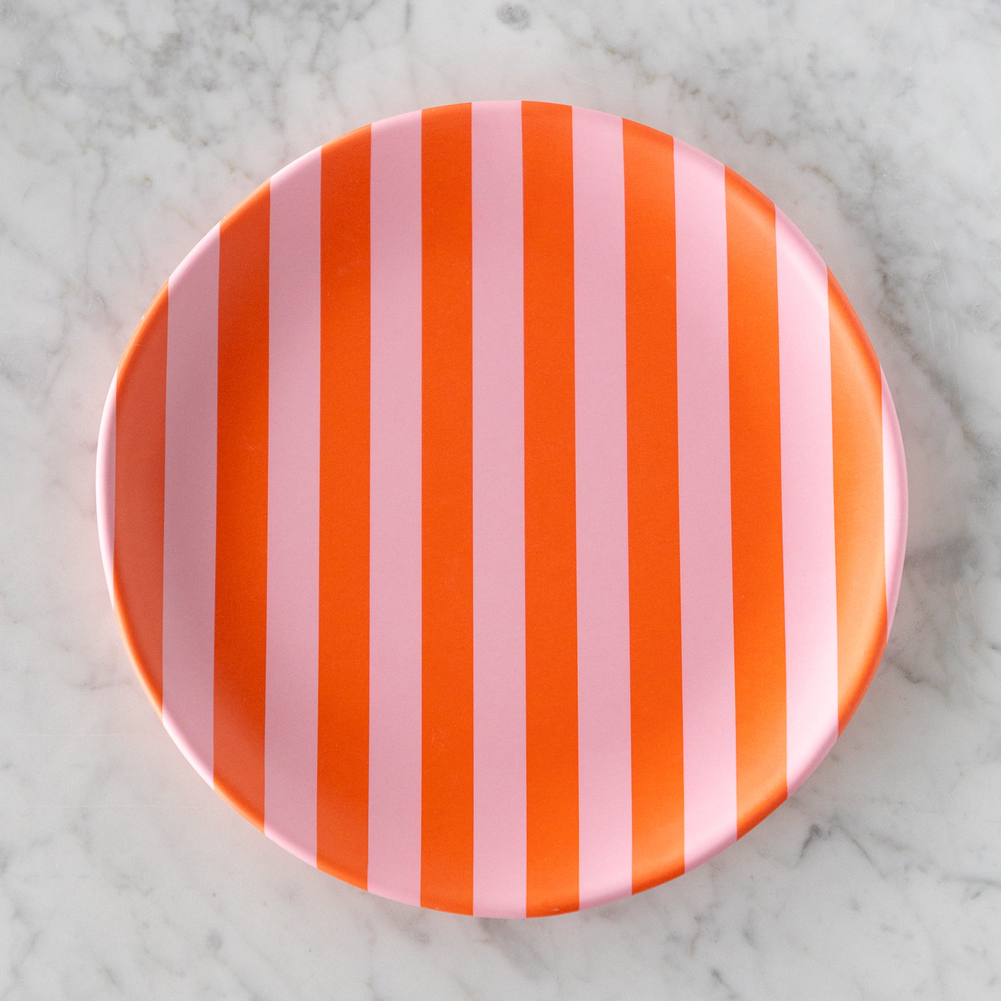 A round Stripe Melamine Plate with alternating vertical pink and orange stripes on a marble table.
