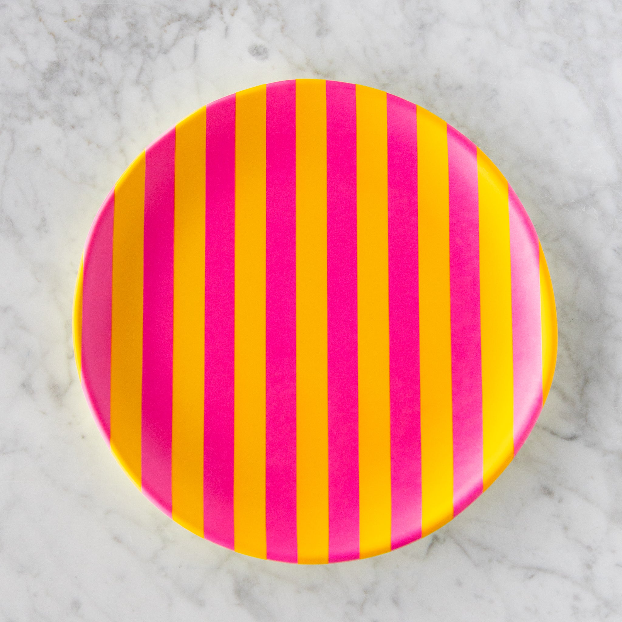 A round Stripe Melamine Plate with alternating vertical hot pink and yellow stripes on a marble table.