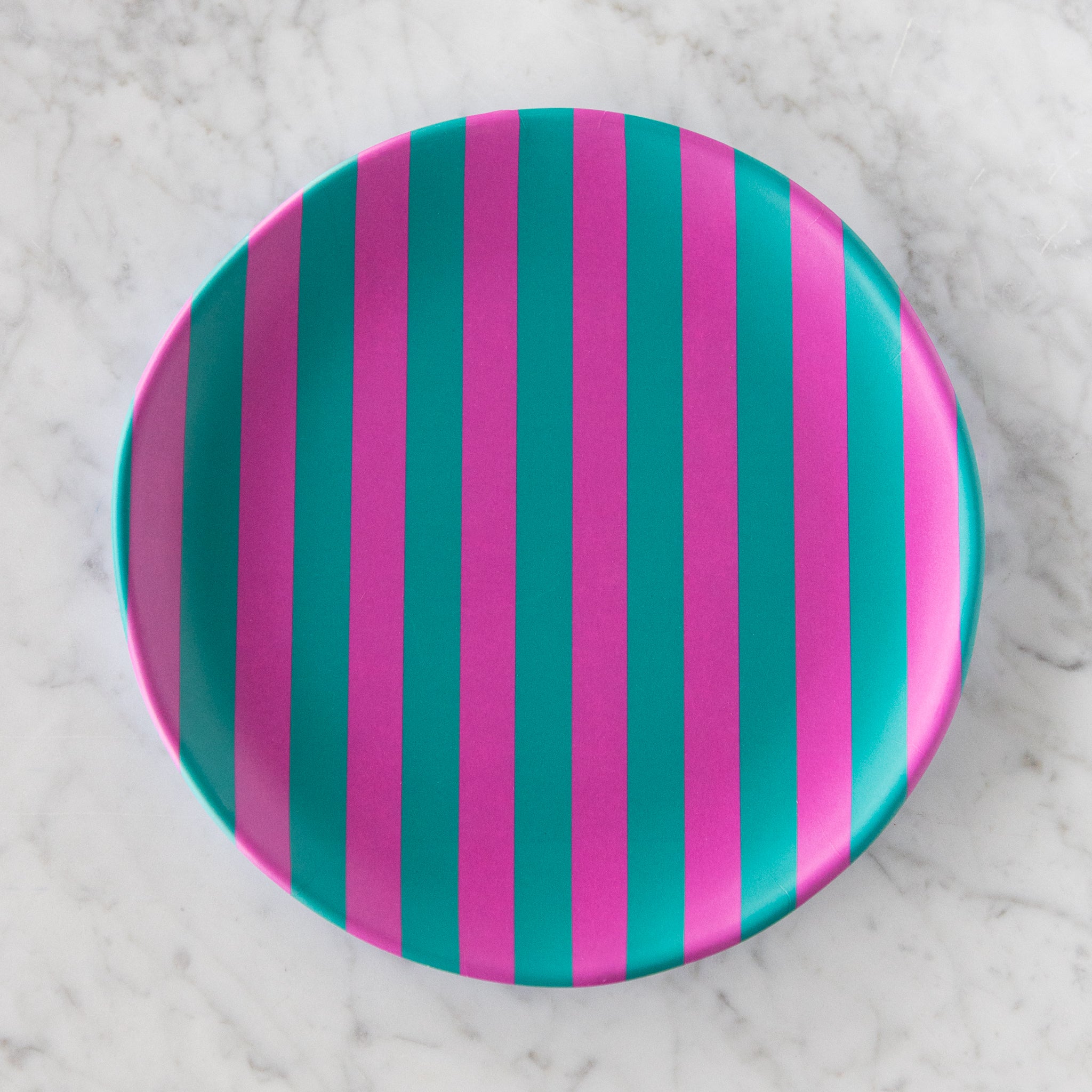 A round Stripe Melamine Plate with alternating vertical fuchsia and turquoise stripes on a marble table.