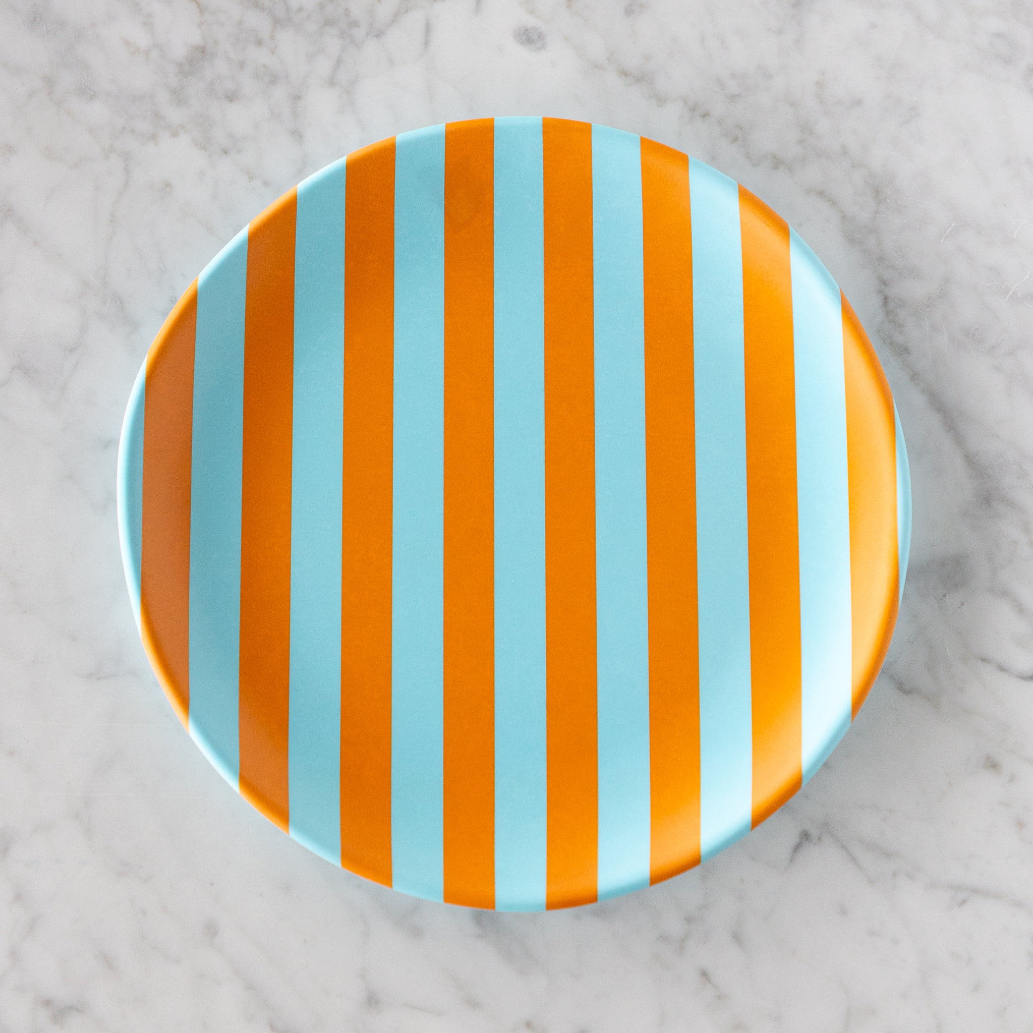 A round Stripe Melamine Plate with alternating vertical light blue and orange stripes on a marble table.