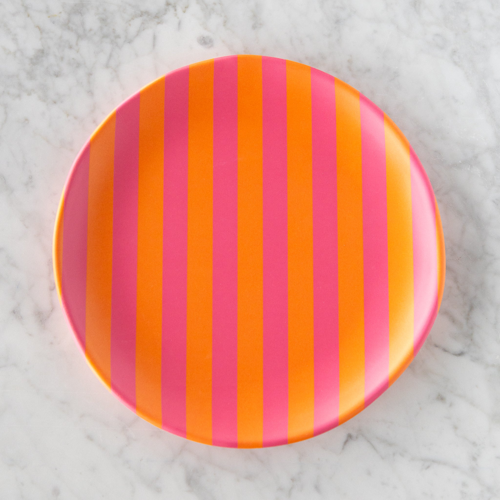 A round Stripe Melamine Plate with alternating vertical pink and orange stripes on a marble table.