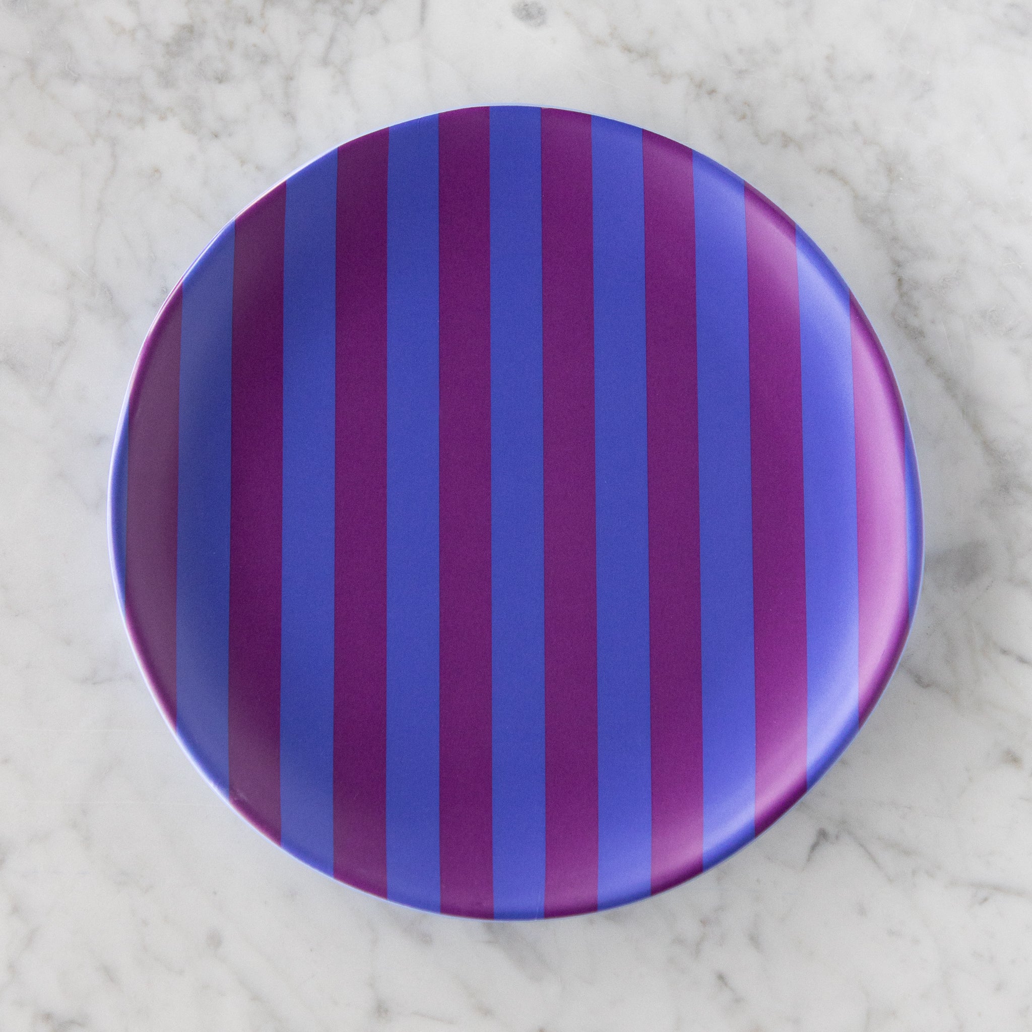 A round Stripe Melamine Plate with alternating vertical purple and blue stripes on a marble table.