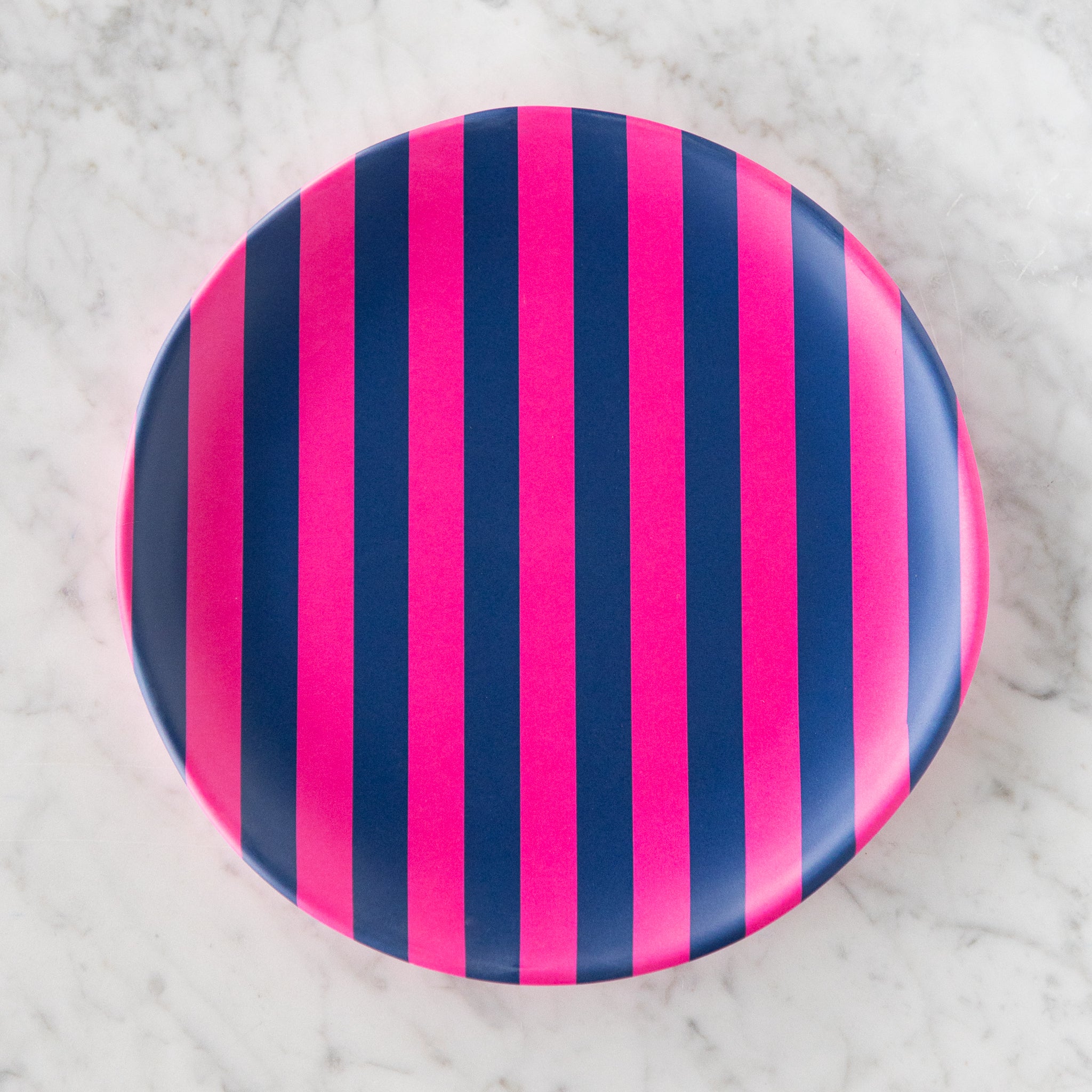A round Stripe Melamine Plate with alternating vertical hot pink and navy stripes on a marble table.