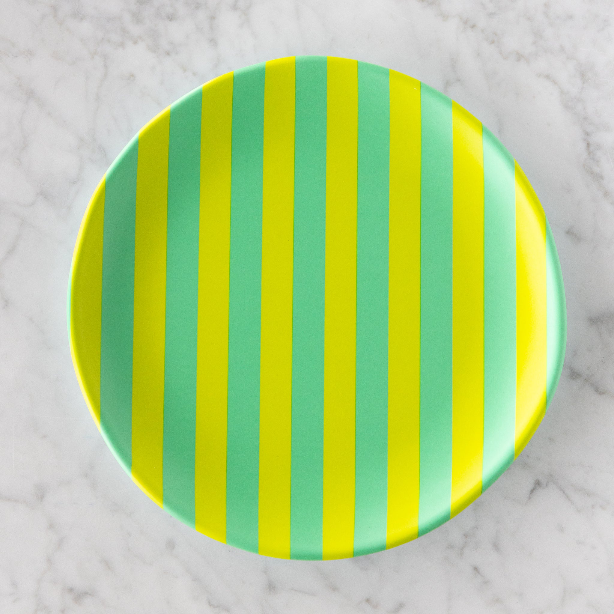 A round Stripe Melamine Plate with alternating vertical lime green and teal stripes on a marble table.