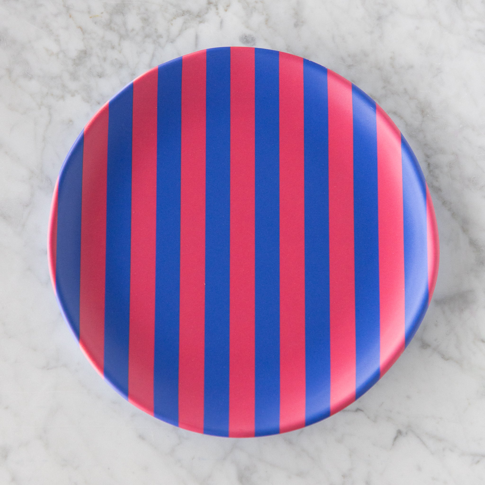 A round Stripe Melamine Plate with alternating vertical pink and blue stripes on a marble table.