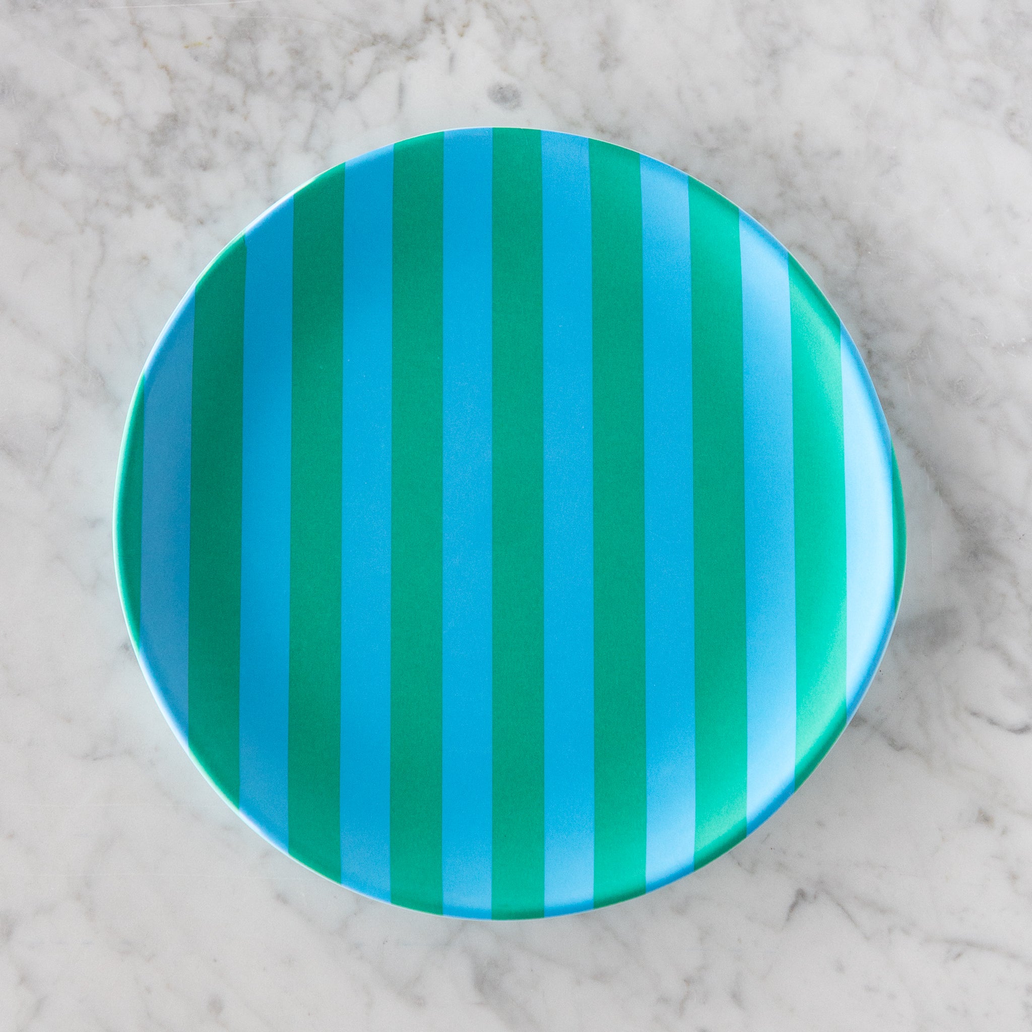A round Stripe Melamine Plate with alternating vertical light blue and turquoise stripes on a marble table.