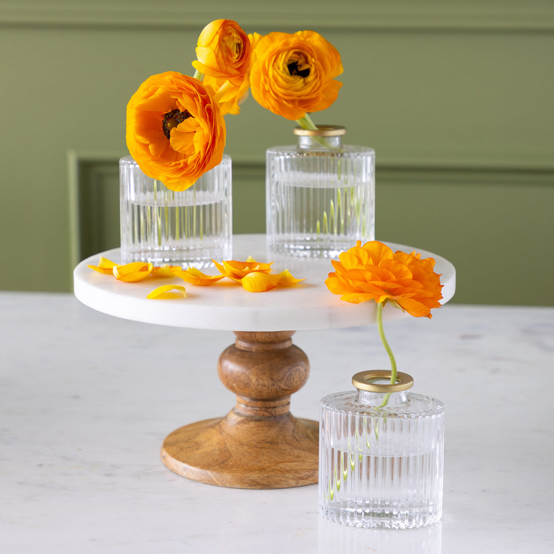 Three Petite Gold Rim Oval Bud Vases  with orange ranunculus flowers and scattered petals are arranged on a round marble and wood cake stand, set against a green wall.