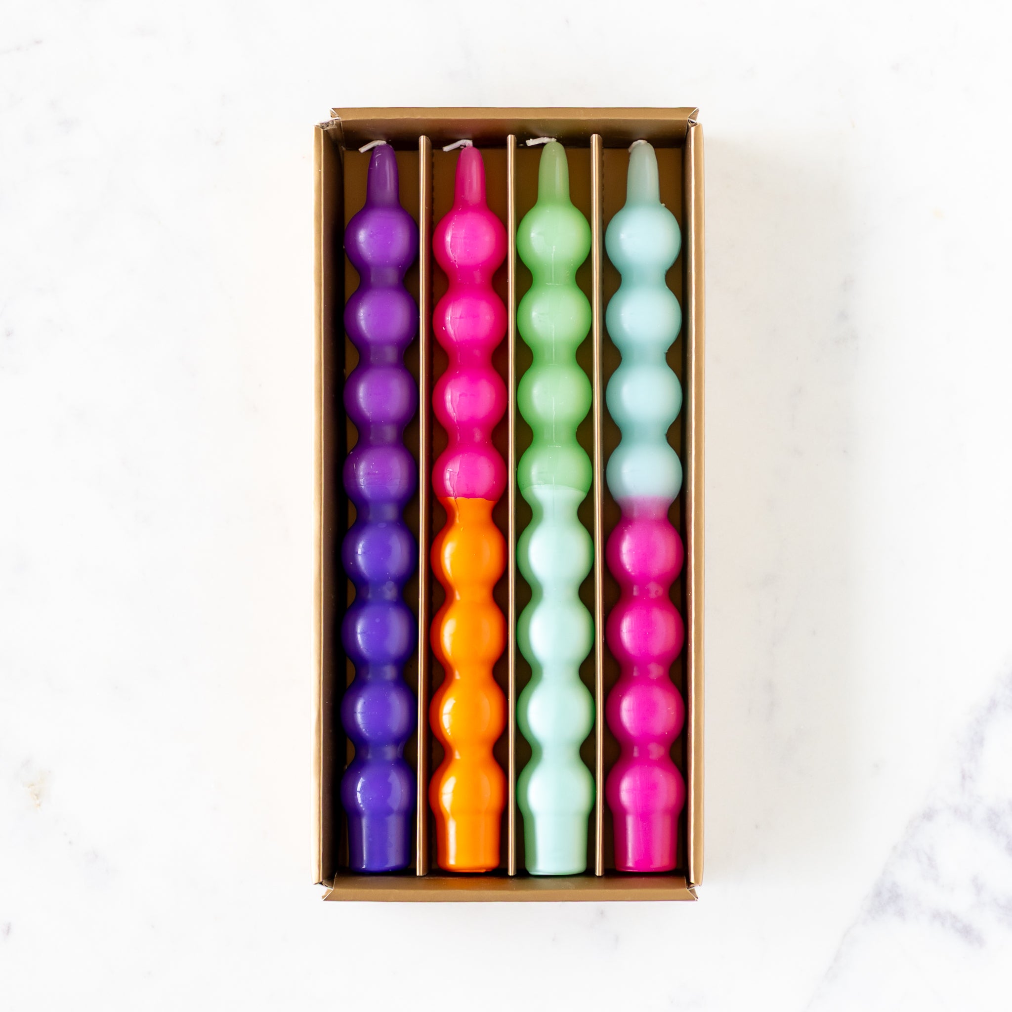 An open box of four Colorful Bubble Taper Candles in two-tone pinks, blues and greens, on a marble table.