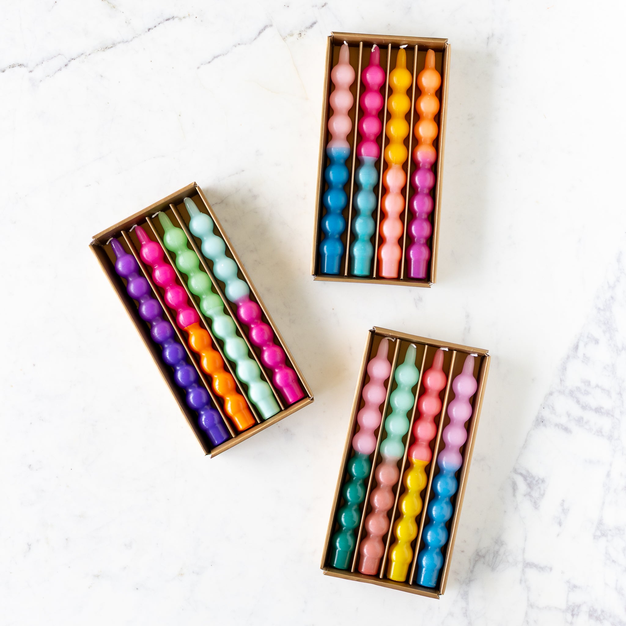 Three open boxes containing sets of 4 colorful Bubble Taper Candles, each twisted and arranged in rows on a white marble surface.