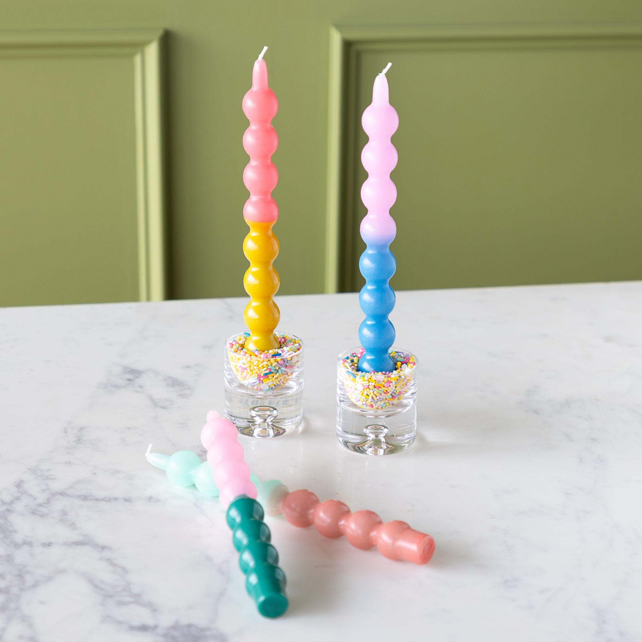 Four Colorful Bubble Taper Candles in pastel hues, two upright in sprinkle-filled glass holders and two lying on a white marble surface.