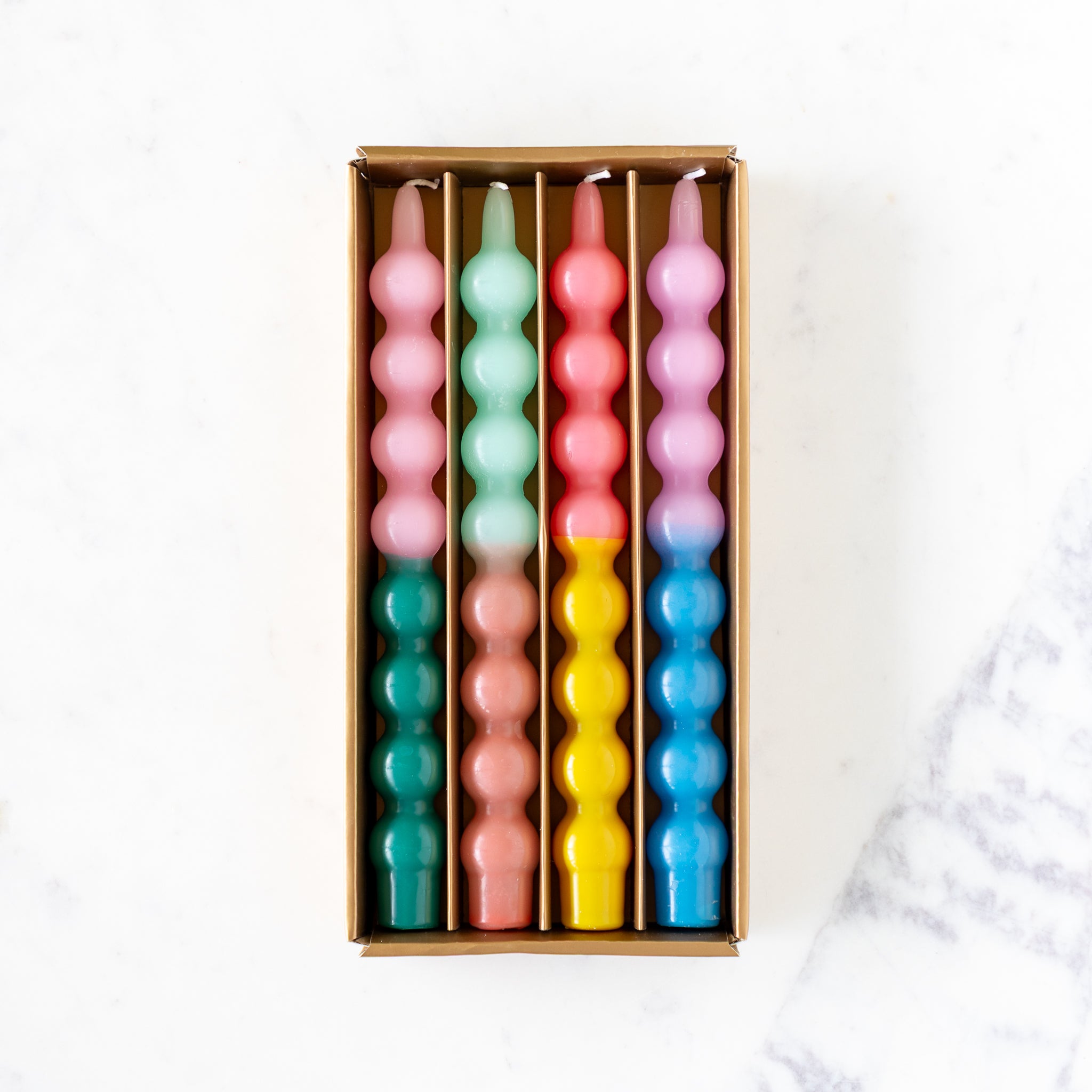 An open box of four Colorful Bubble Taper Candles in two-tone pastel colors, on a marble table.
