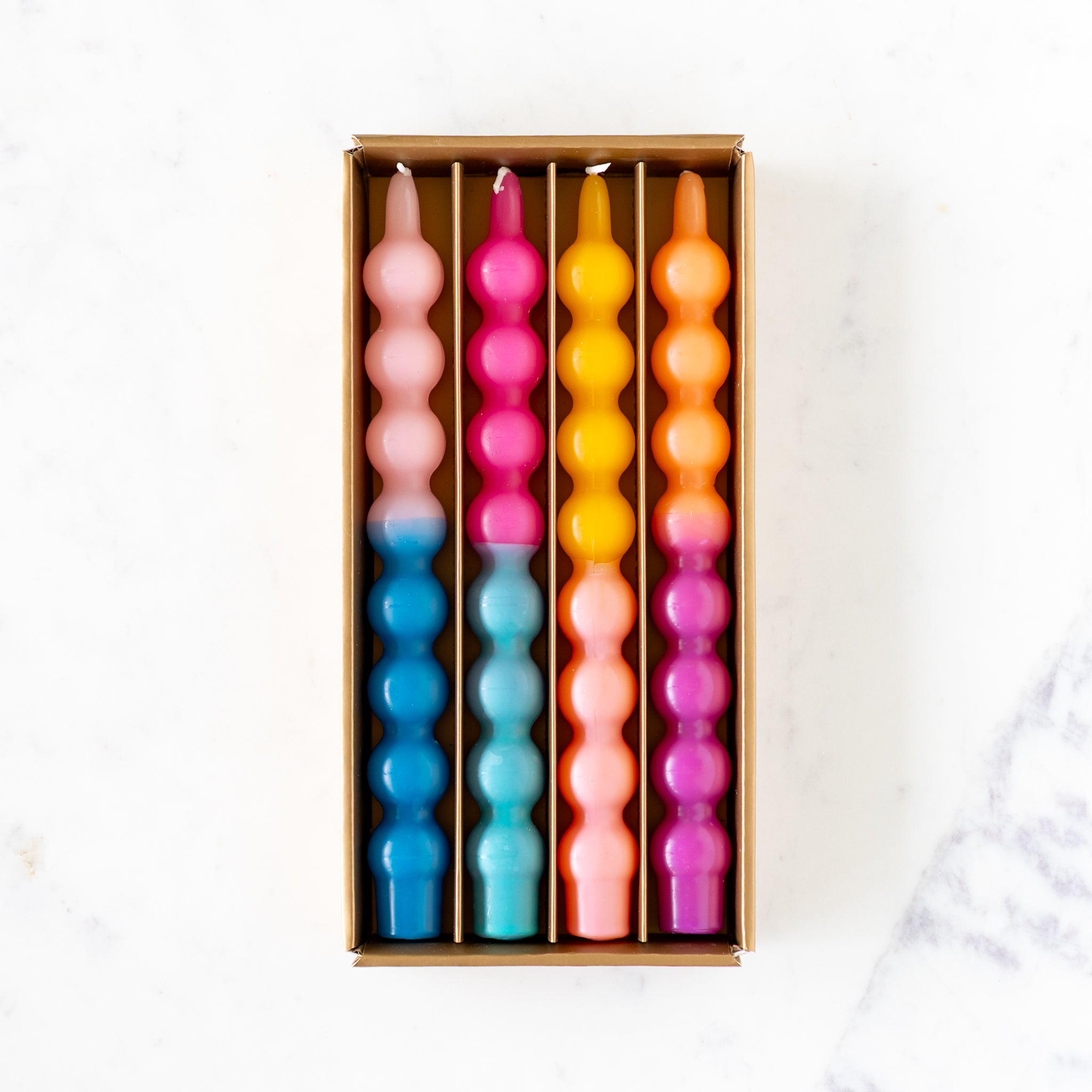 An open box of four Colorful Bubble Taper Candles in two-tone blues, pinks and yellows, on a marble table.