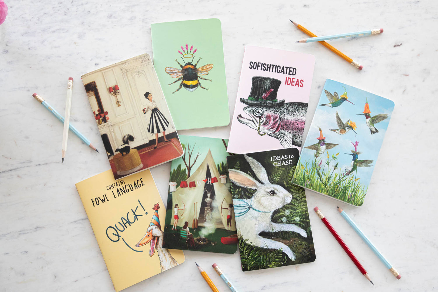 Hester &amp; Cook Notebooks featuring artwork by Vicki Sawyer, Elizabeth Foster, Janet Hill and Bewilderbeest, scattered on a marble table surrounded by pencils.