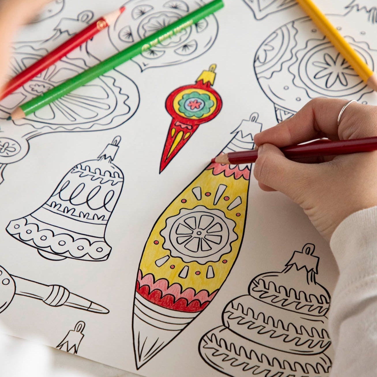 A woman coloring an ornament on the Christmas Coloring Placemat with scattered colored pencils.