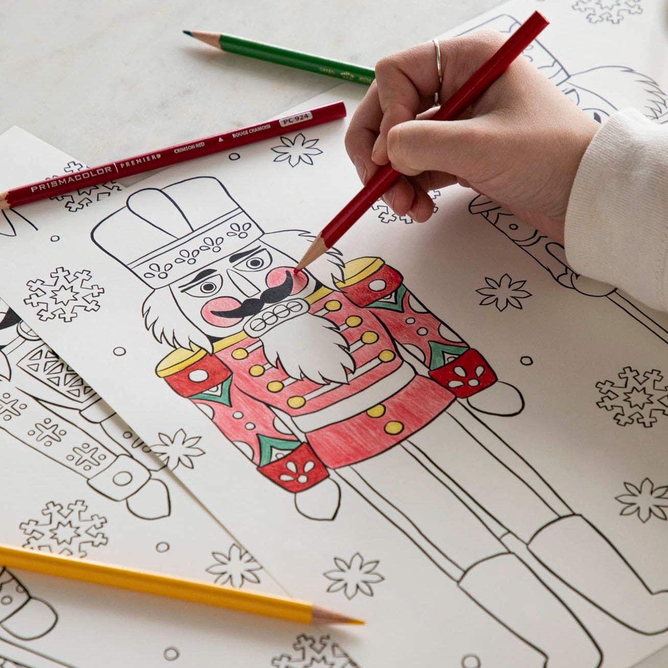 A woman coloring a nutcracker on the Christmas Coloring Placemat with scattered colored pencils.